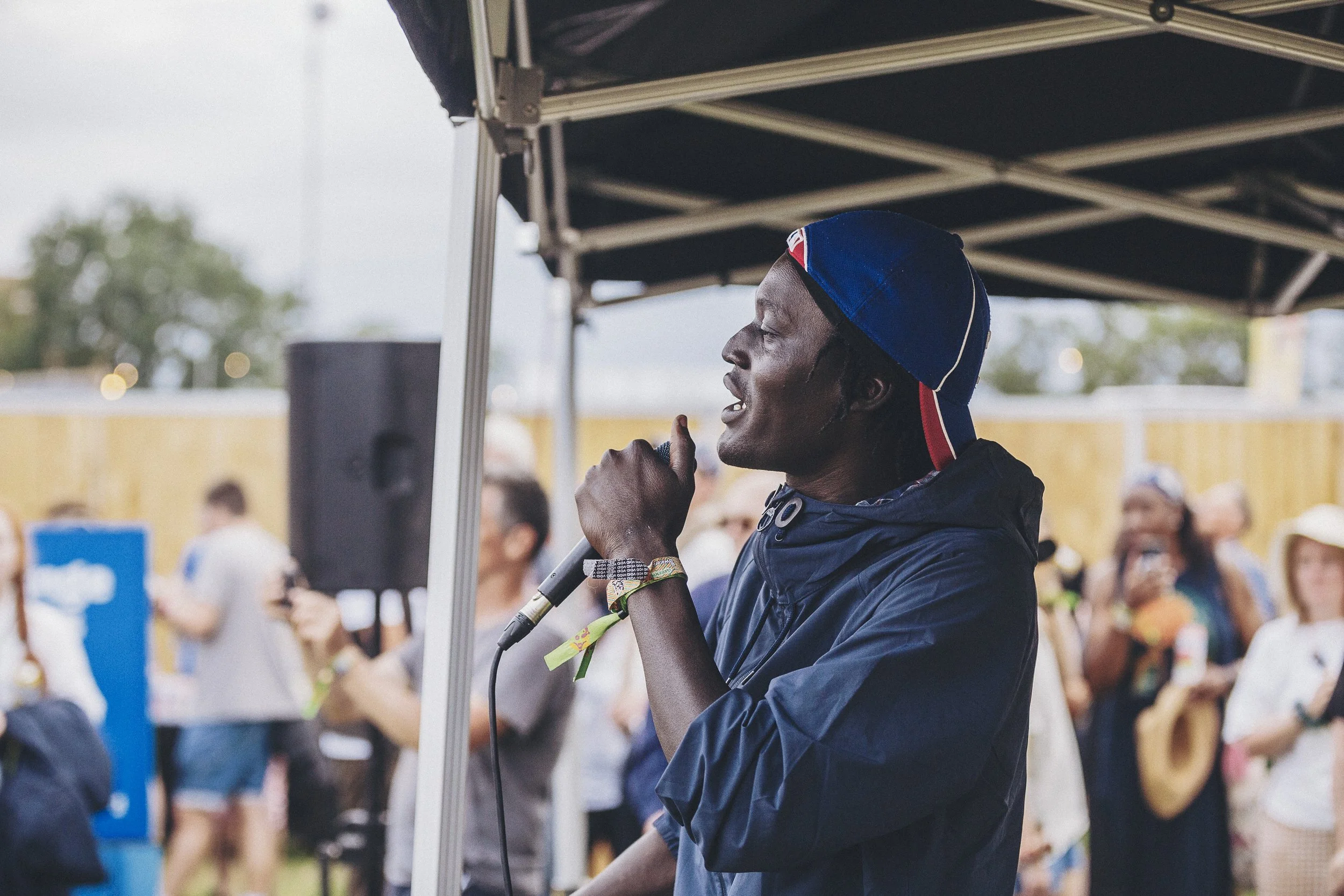 A person speaking into a microphone at an outdoor event, with a crowd in the background.