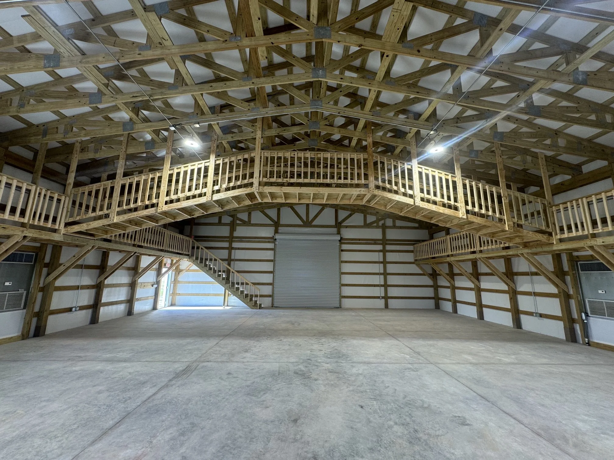 Interior of a large building with unfinished wooden framework and a high ceiling, featuring balcony railings and an open roller door at the far end.