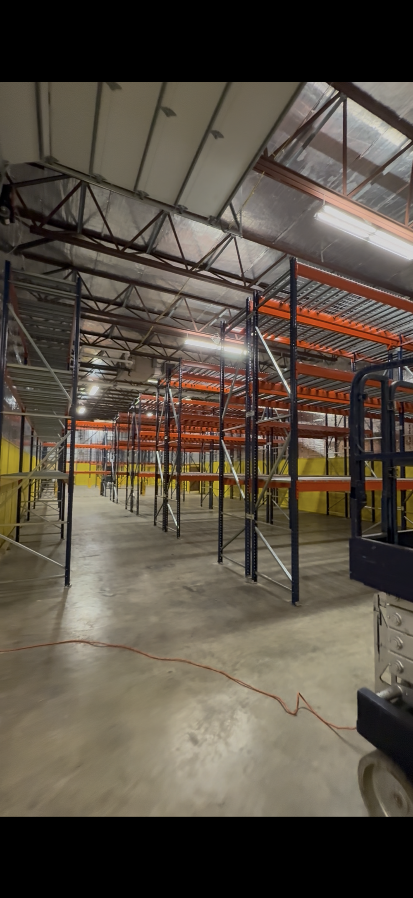 Empty warehouse with metal shelving units and scaffolding, construction equipment, and overhead lighting.