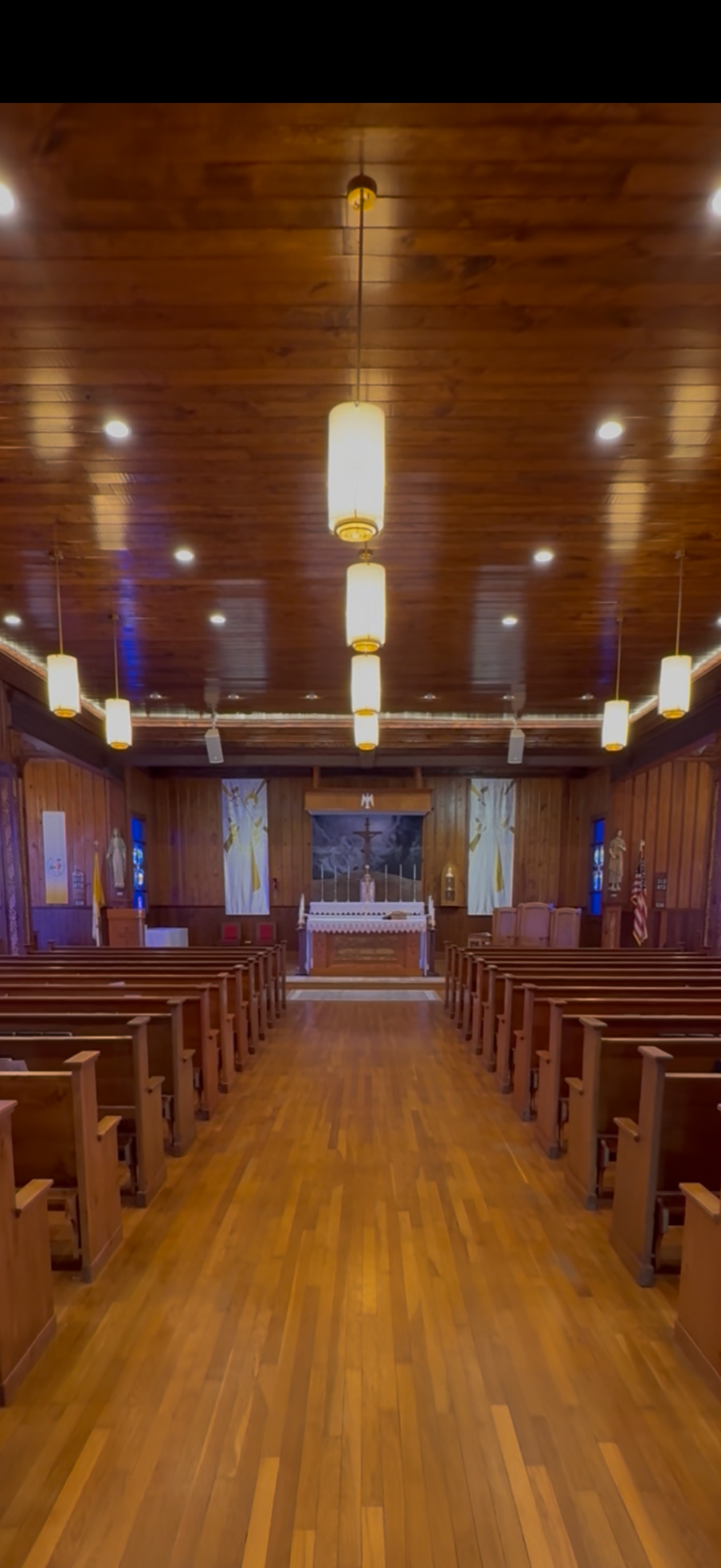 Interior of a small Christian chapel with wooden pews, a wooden altar with a crucifix, candles, and religious statues, lit by hanging pendant lights and wall lamps.