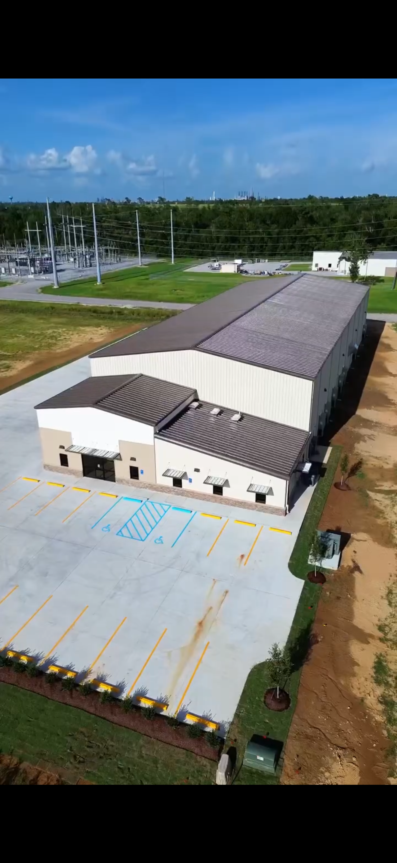 An aerial view of a large commercial building with a parking lot that has two accessible parking spaces painted in blue. The building has a white exterior with a metal roof, and surrounding it are landscaped areas with trees and grass, along with a dirt path.