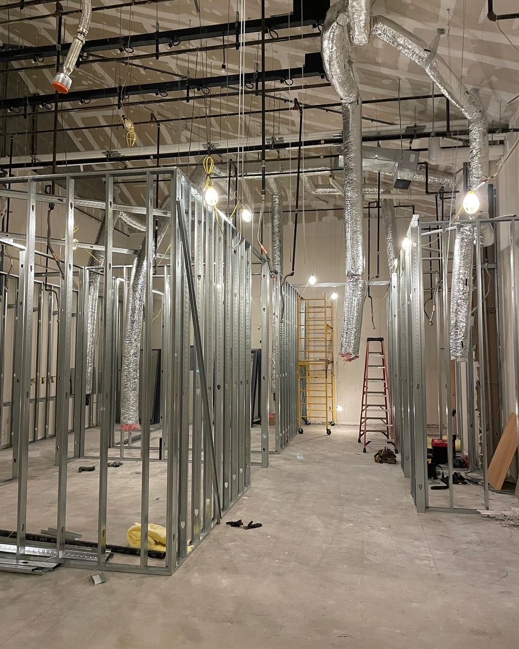Construction site with metal framing, exposed ductwork, and hanging light bulbs, indicating an interior renovation.