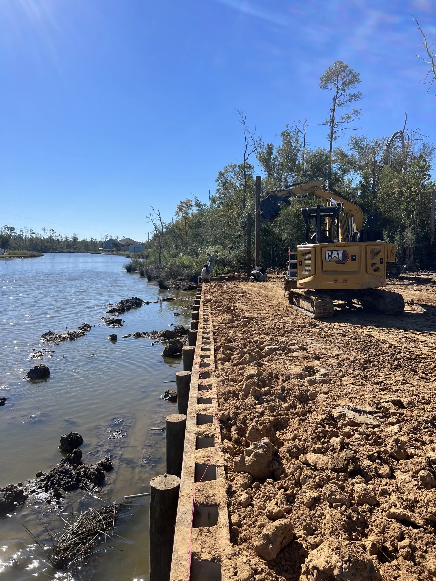 Construction work along the water's edge with a yellow Cat excavator and workers installing wooden posts for a shoreline barrier, with trees and houses in the background under a clear blue sky.