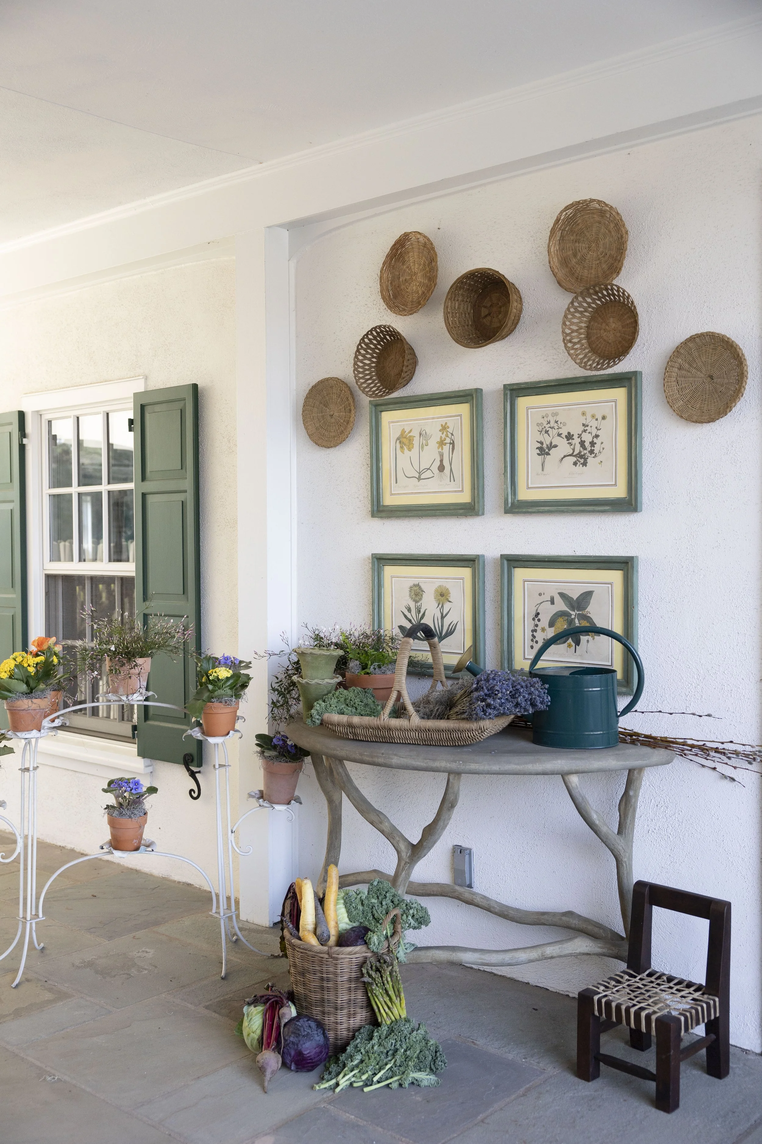 Outdoor scene with a white textured wall decorated with a cluster of woven baskets and framed botanical prints. A table with potted flowers, a basket of vegetables, and gardening tools is in the scene, along with garden-themed decor and a window with green shutters.