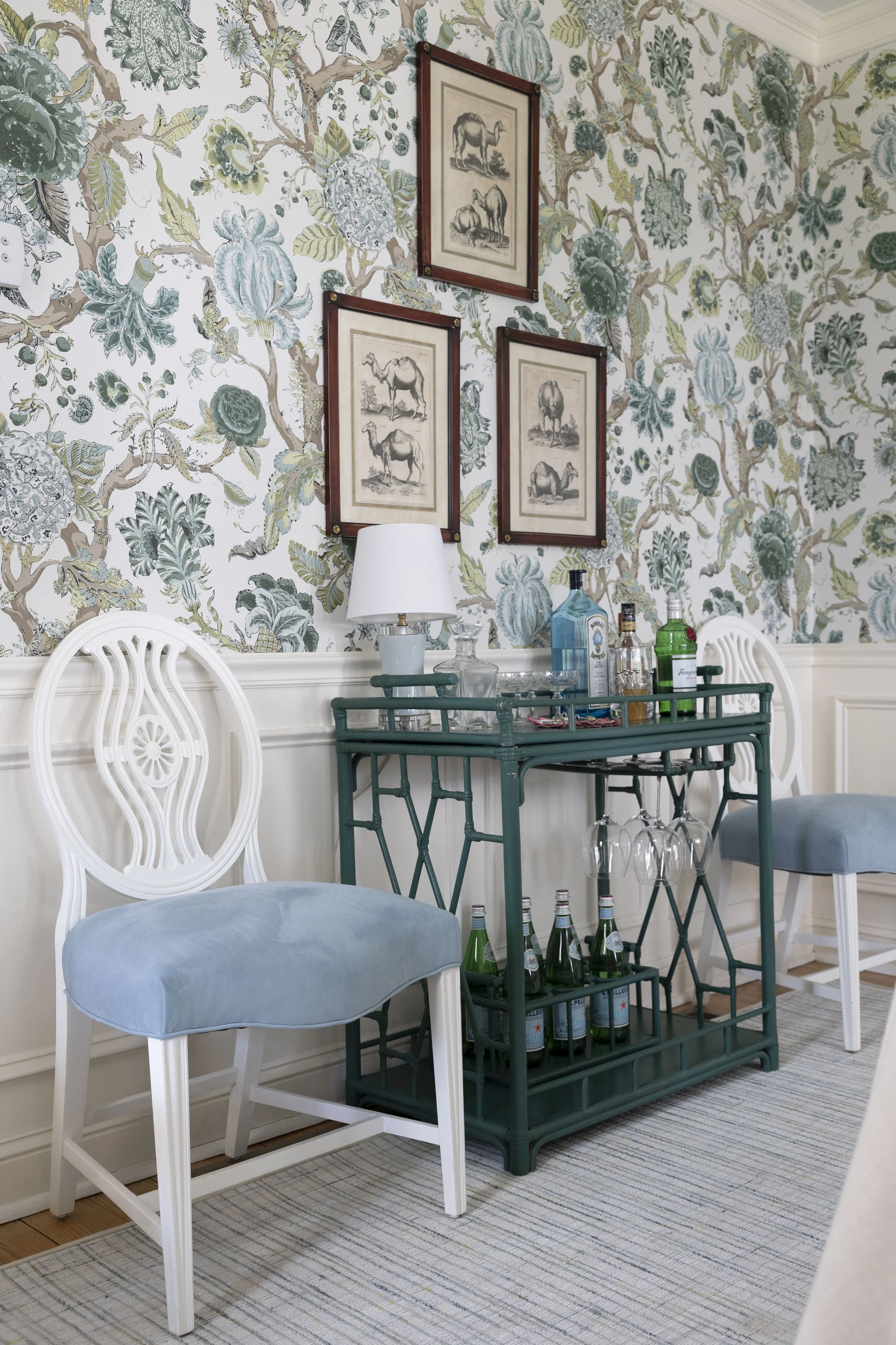 A decorated dining room corner with a patterned wallpaper featuring blue and green foliage and animals. A white chair with a light blue cushioned seat and a green metal bar cart with bottles and glassware are in the foreground. Framed saddle and camel sketches hang on the wall, and there is a table lamp on a sideboard.