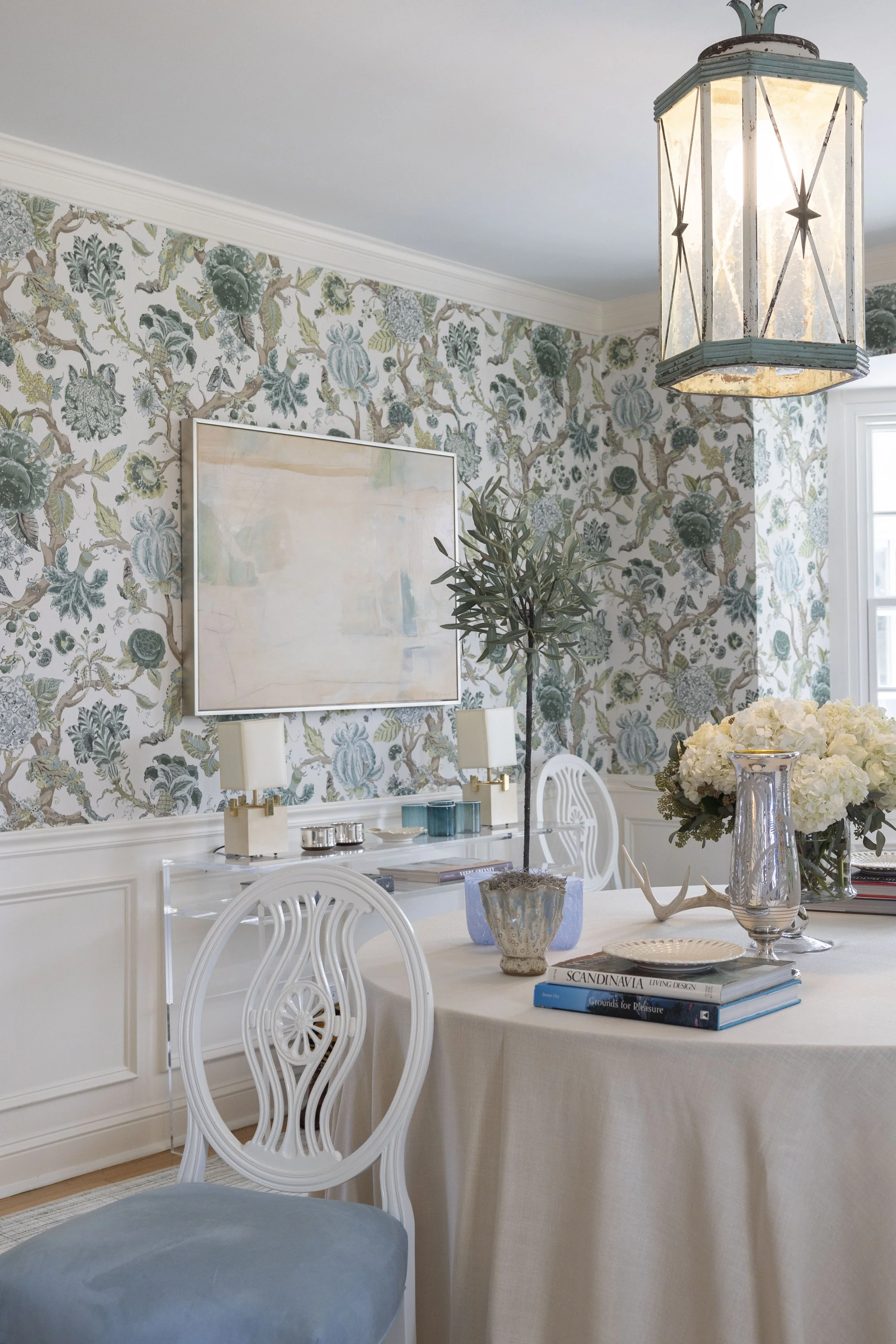 A dining room with floral wallpaper, a white table with books and decor, a large silver vase with white flowers, a smaller decorative bowl, a blue velvet cushioned chair, a wooden chair, a chandelier, and window light.