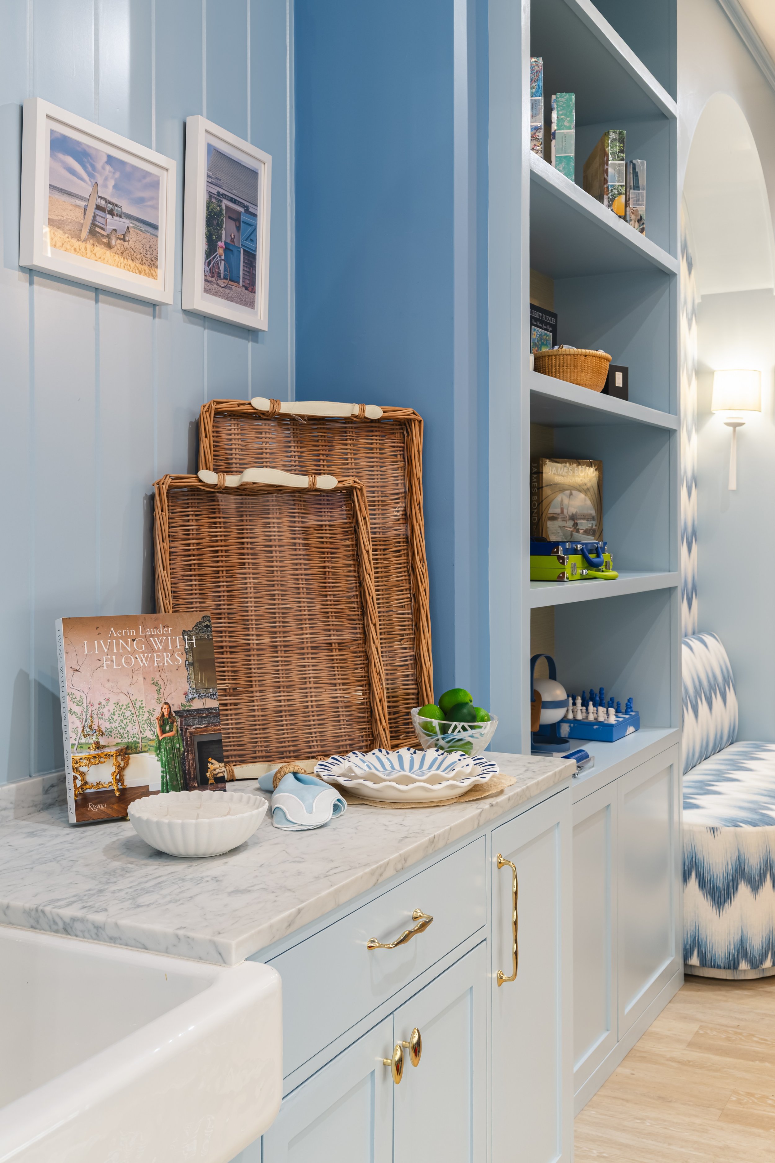 A kitchen or dining area with light blue cabinets with gold handles, a marble countertop, and some decorative items such as bowls, books, and plants. There are two framed beach-themed photos on the wall, a wicker tray, and a small bowl of green apples. A pitcher with a lid and a blue and white striped curtain are also visible.