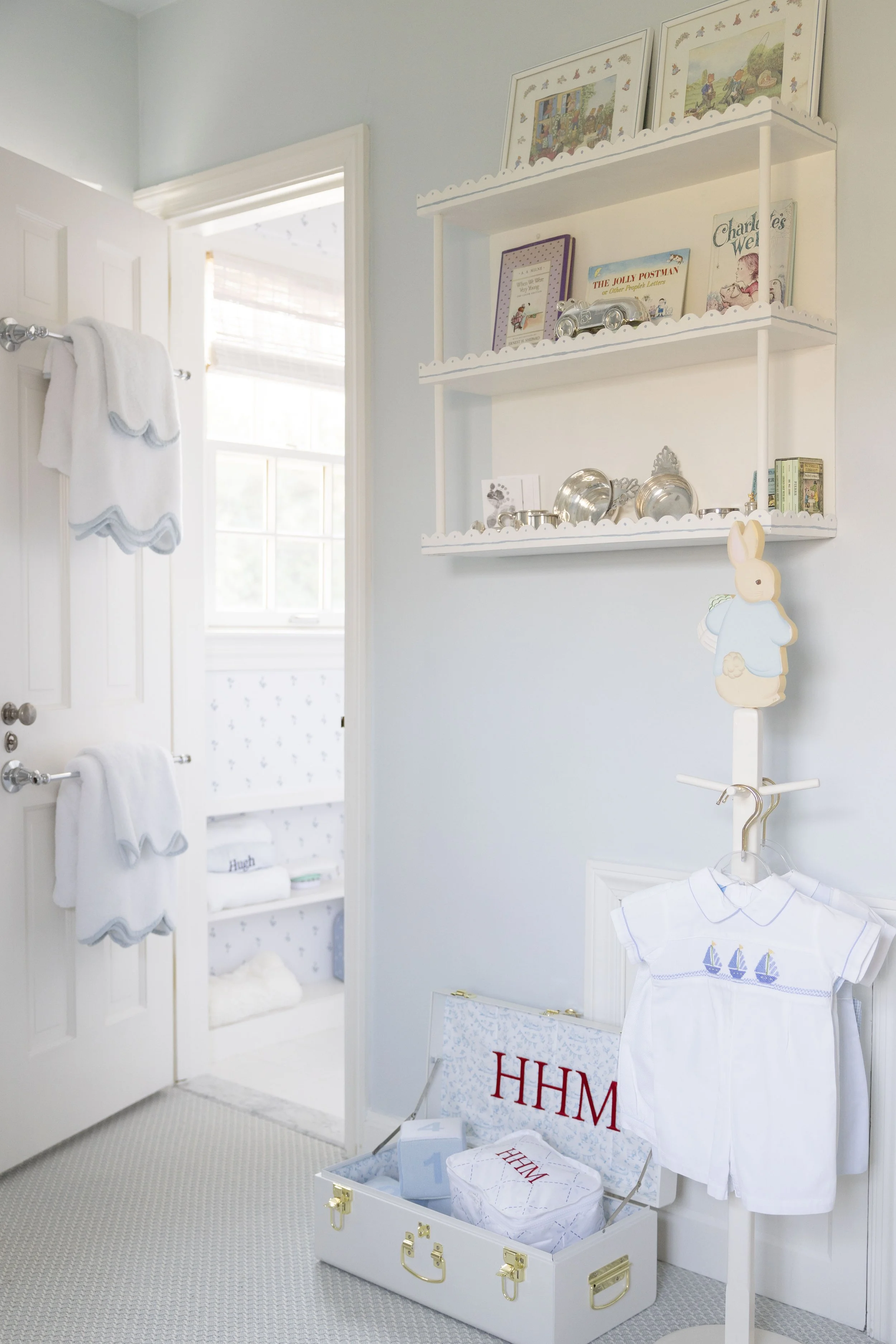 A nursery room decorated with blue and white colors, featuring a white open wardrobe with folded clothes, a white bookshelf with books and decorative items, a white coat rack with children's clothes, and soft lighting.