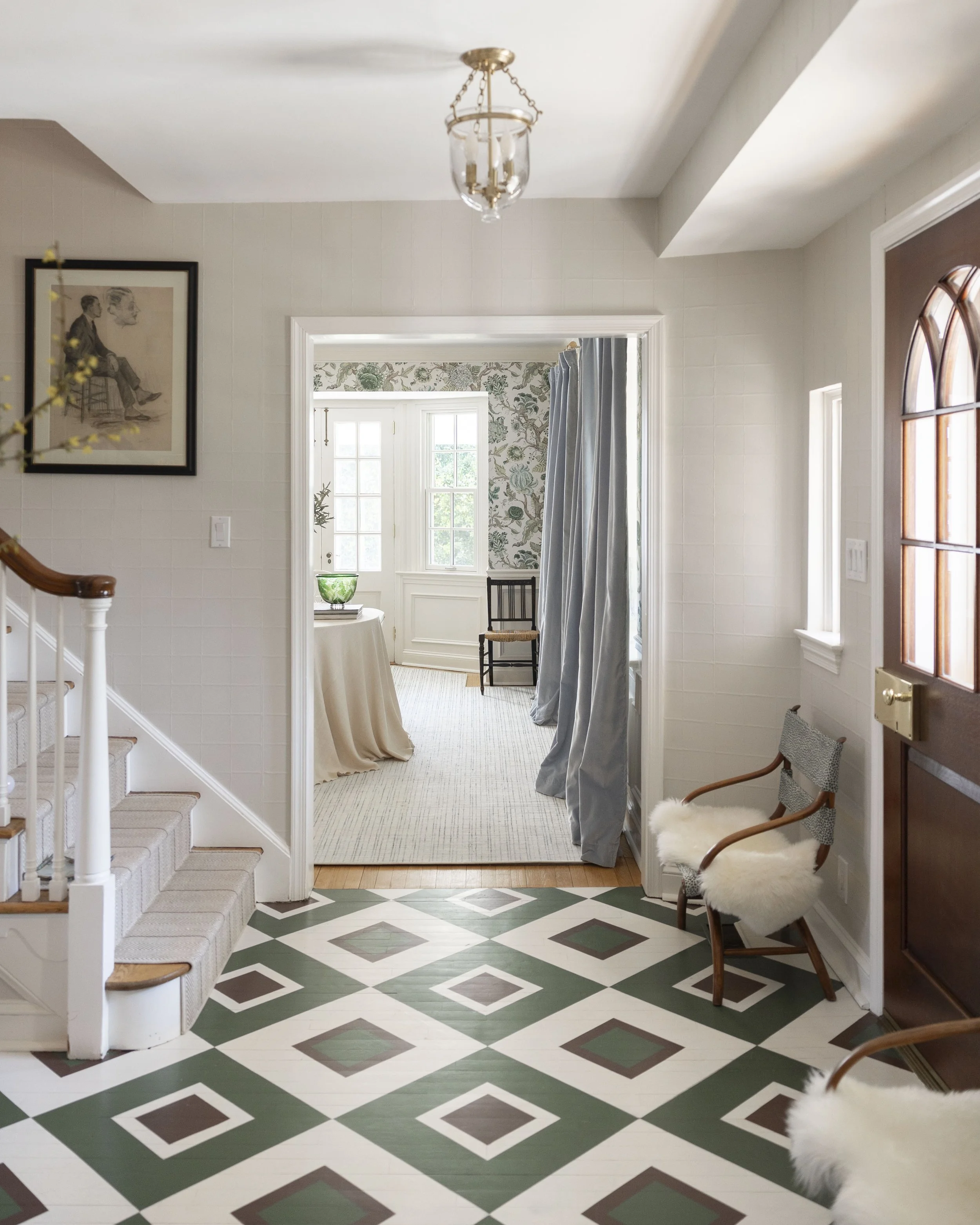 Entryway with patterned tile floor, wooden staircase, white walls, framed artwork, and a chair with a white fur cushion.