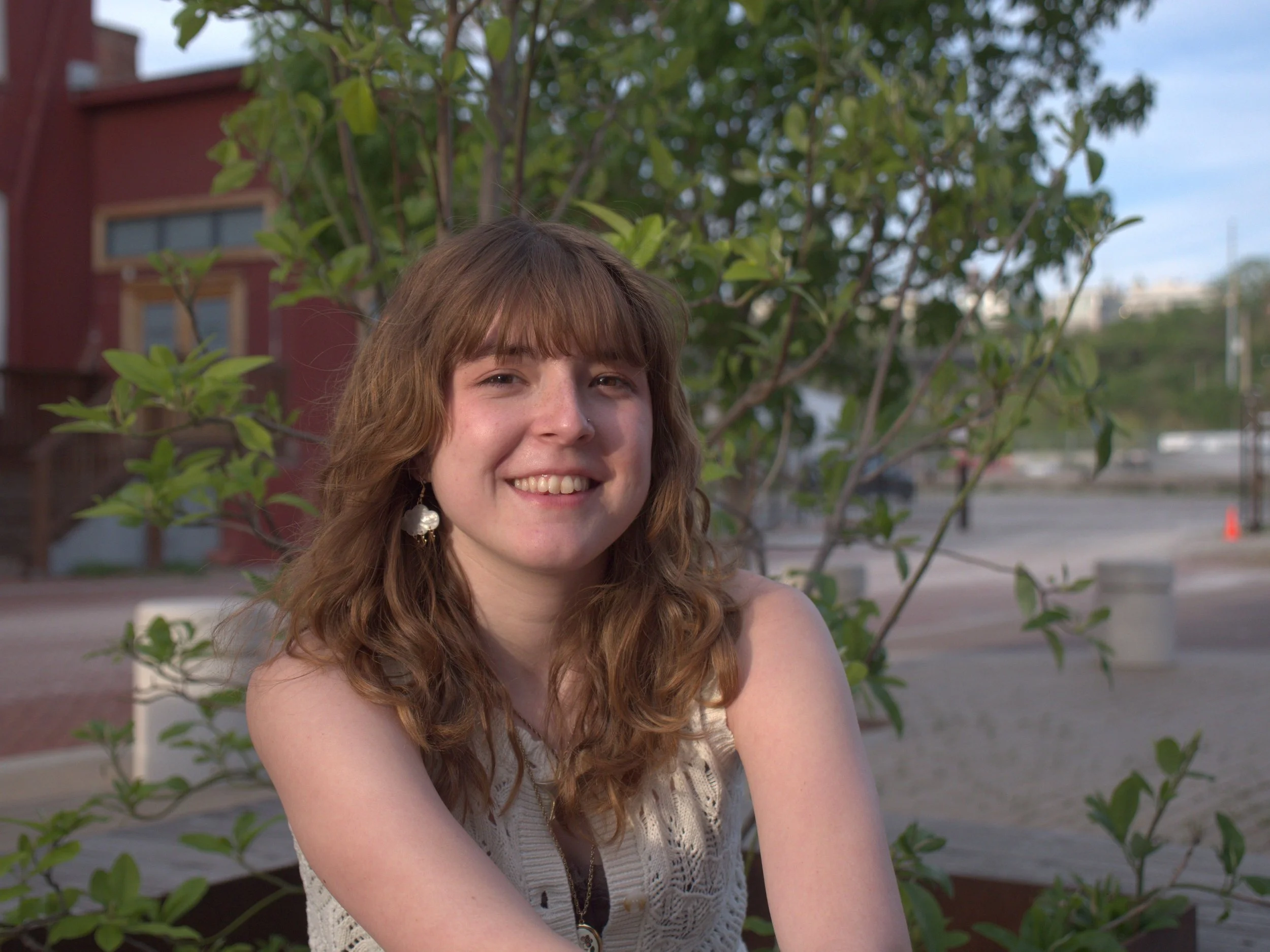A young woman with long curly brown hair smiling outdoors with a tree and building in the background. Ivy England a professional Illustrator.