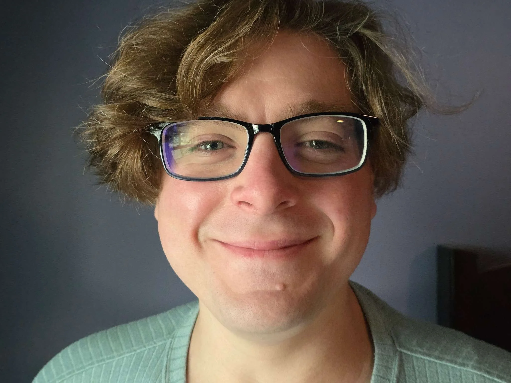 Close-up photo of a person with wavy blonde hair, glasses, and a pleasant expression, wearing a greenish top. The background is dark. David Hunter a professional children's book author.