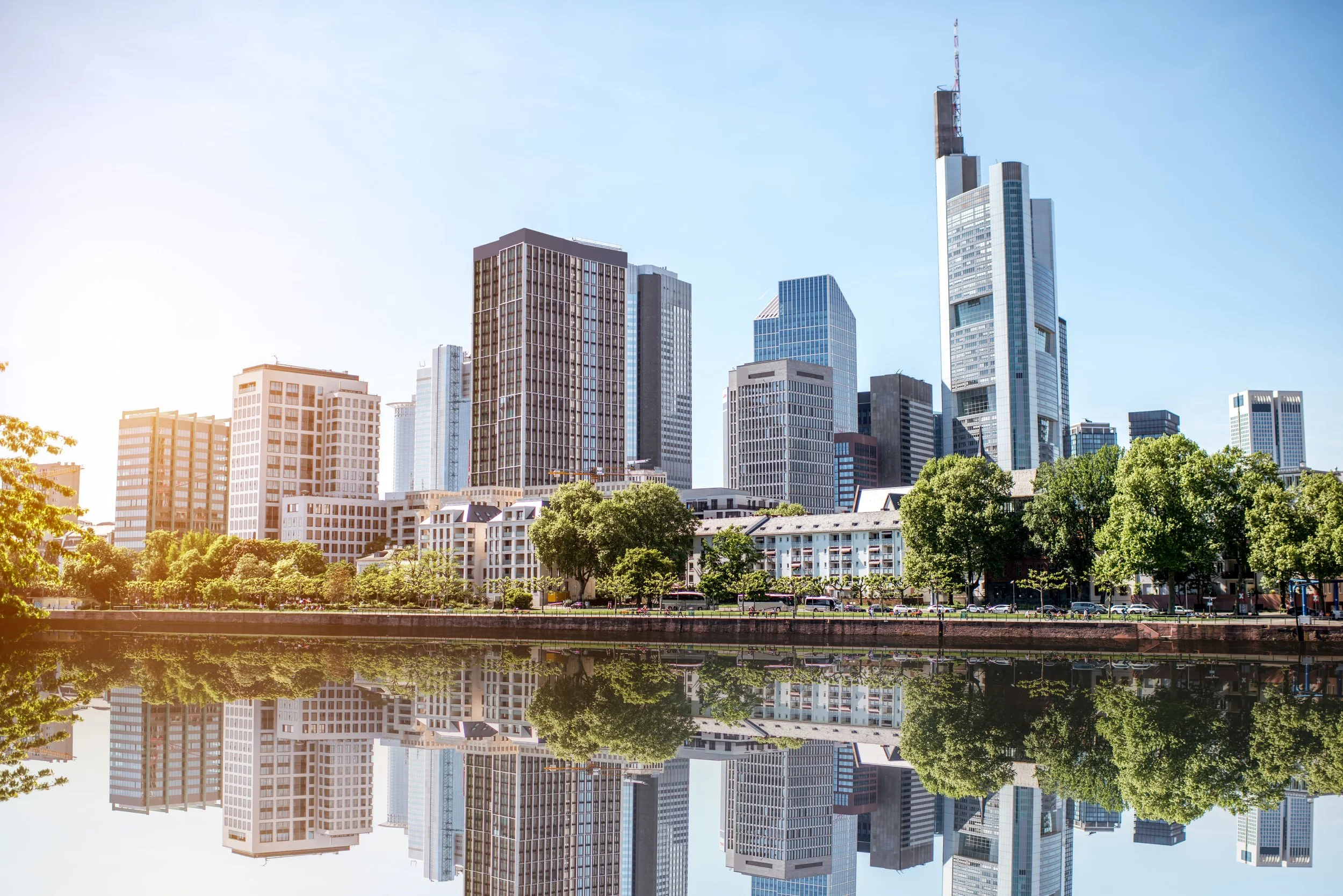 Skyscrapers and high-rise buildings in a city skyline, reflected in a river with green trees in the foreground, under a clear blue sky.