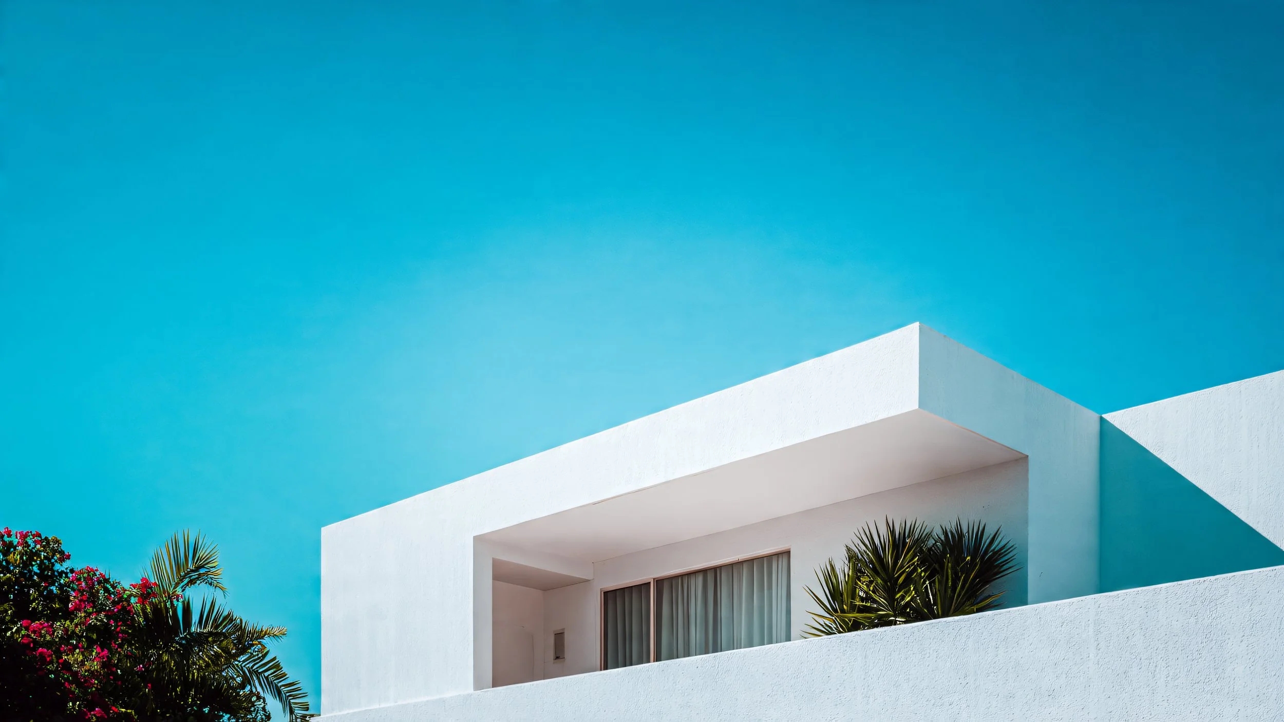 White modern building with clean lines, set against a clear blue sky, surrounded by greenery including palm trees and flowering bushes.