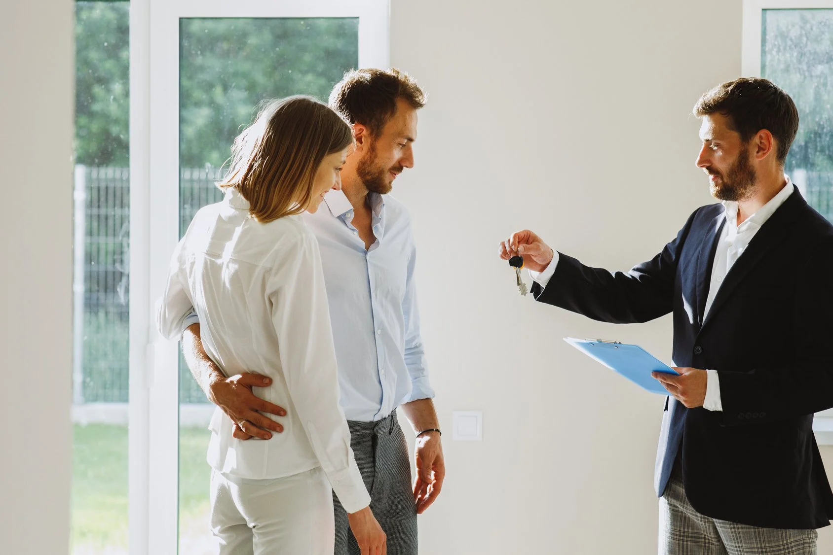 A couple receives house keys from a real estate agent indoors, near large windows showing a garden outside.