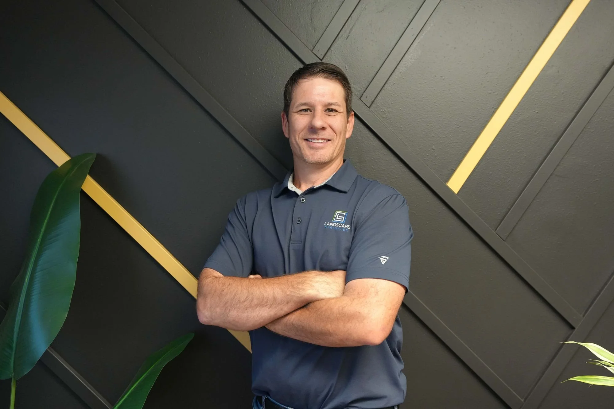 A man with short brown hair, wearing a dark gray polo shirt with a small logo on the chest and crossed arms, standing in front of a dark wall with diagonal yellow accents and large green leaves on the left side of the image.