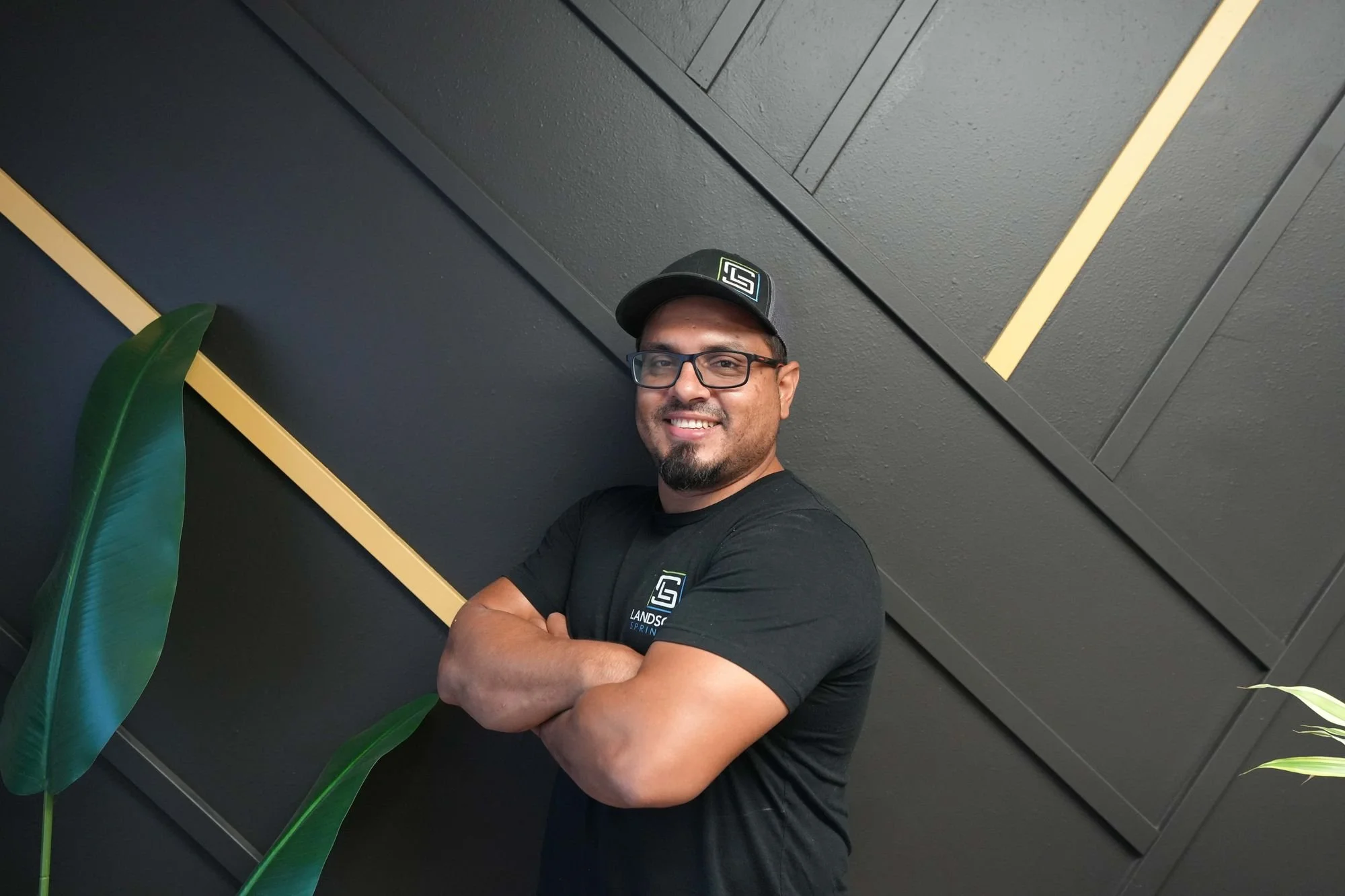 A smiling man wearing glasses and a black cap standing with his arms crossed in front of a dark wall with yellow accents and large green plants.