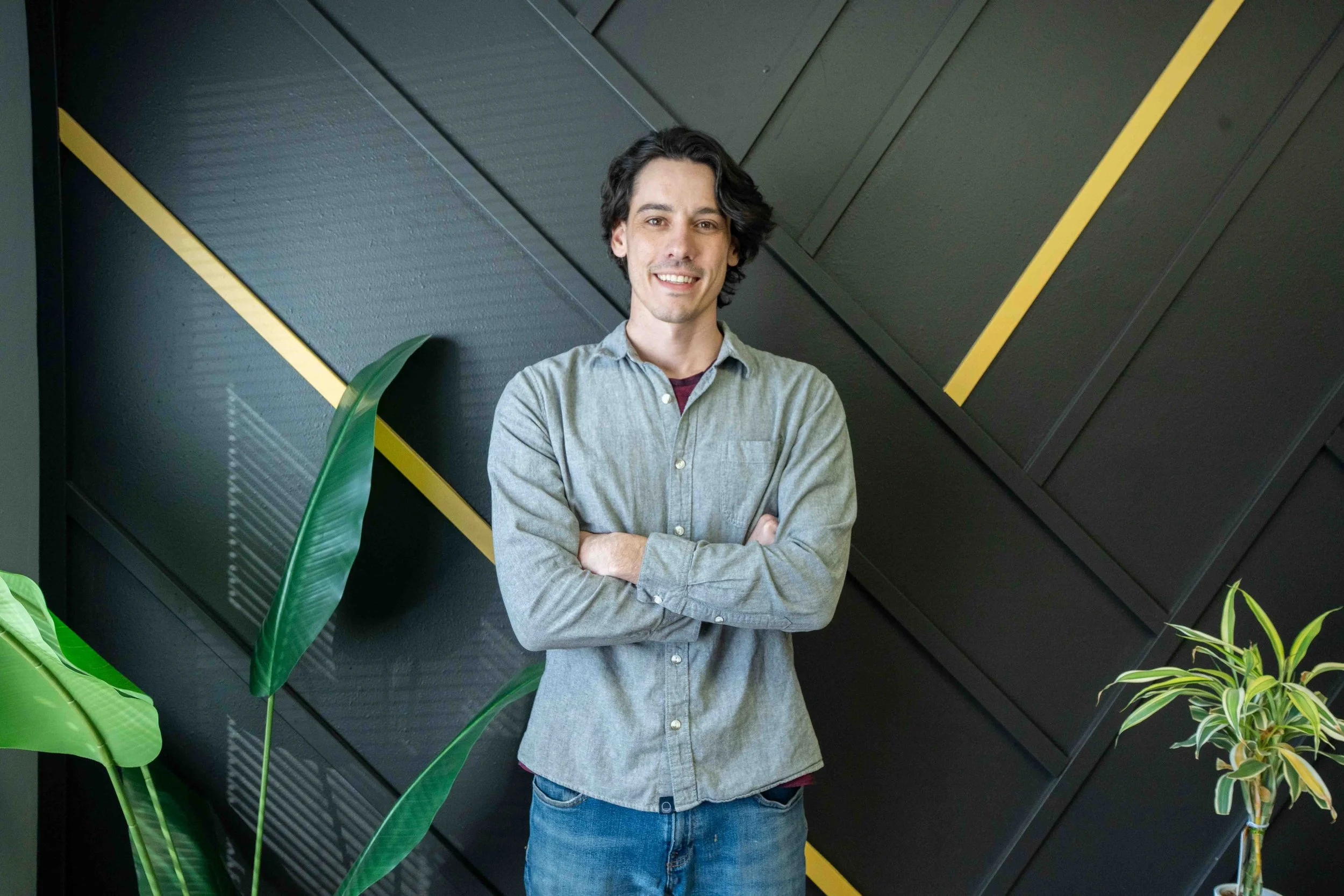 A young man with dark, wavy hair and light skin smiling with arms crossed, standing indoors against a wall with black textured panels accented with yellow lines, near large green leafy plants.