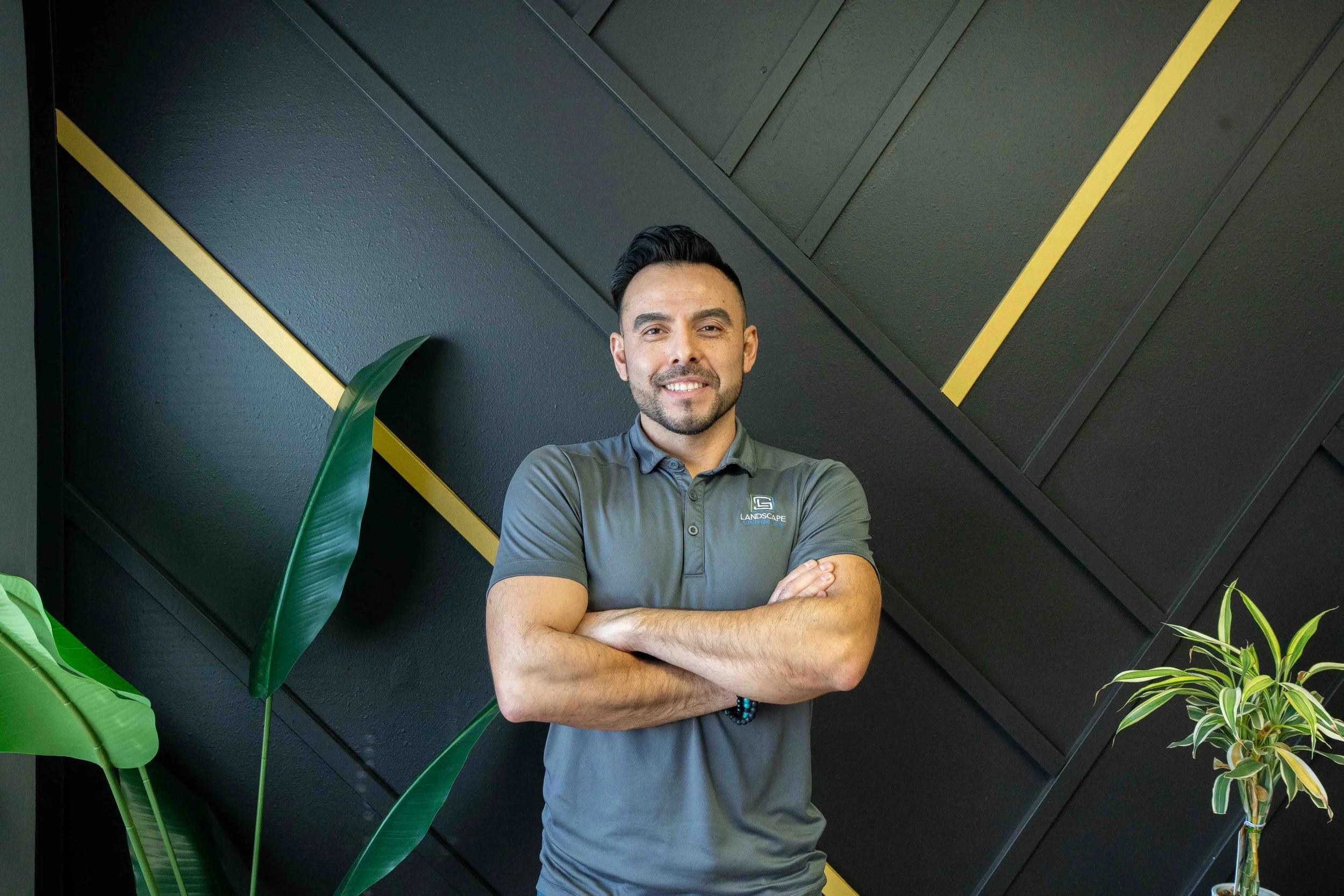 A man with crossed arms smiling in front of a black wall with diagonal yellow lines, surrounded by green plants.