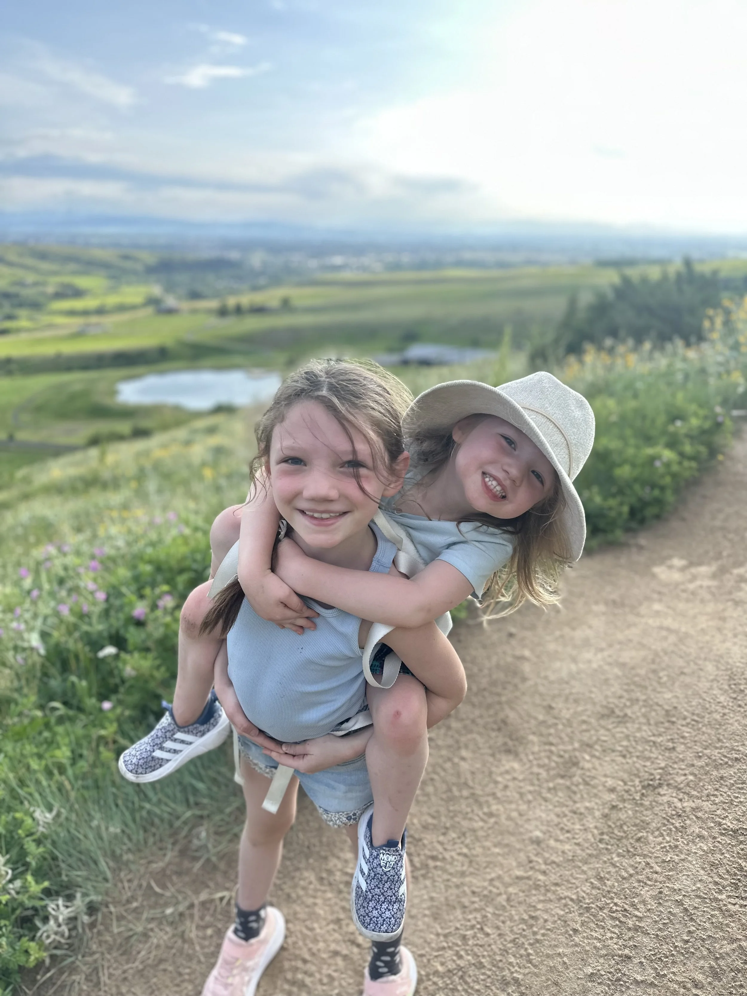 Two young children happily playing outdoors on a dirt trail with green fields and a pond in the background, smiling and wearing casual clothes and hats.