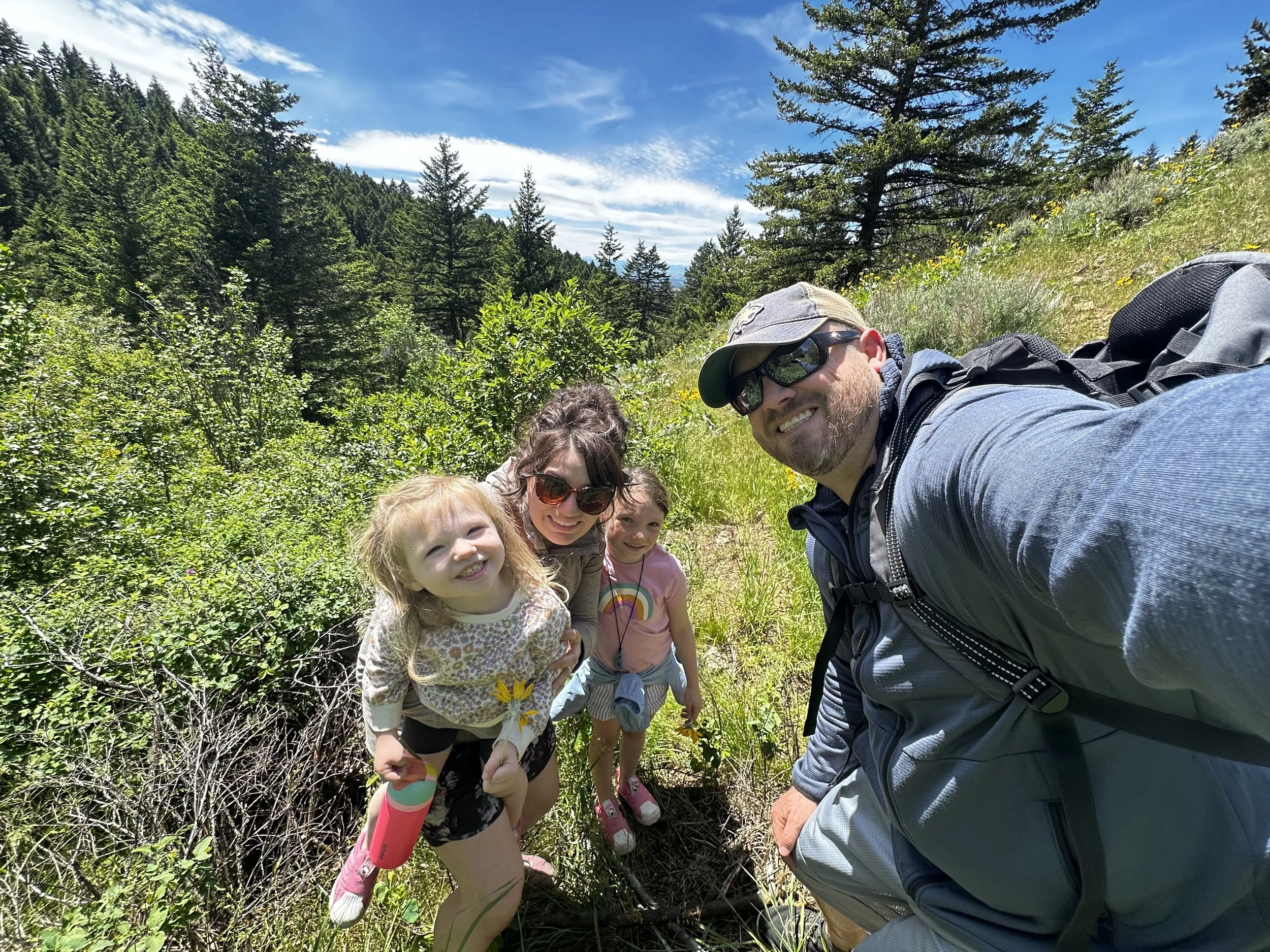 A group of children and an adult taking a selfie outdoors in a forest with tall trees and blue sky. The children are smiling and holding small yellow flowers.