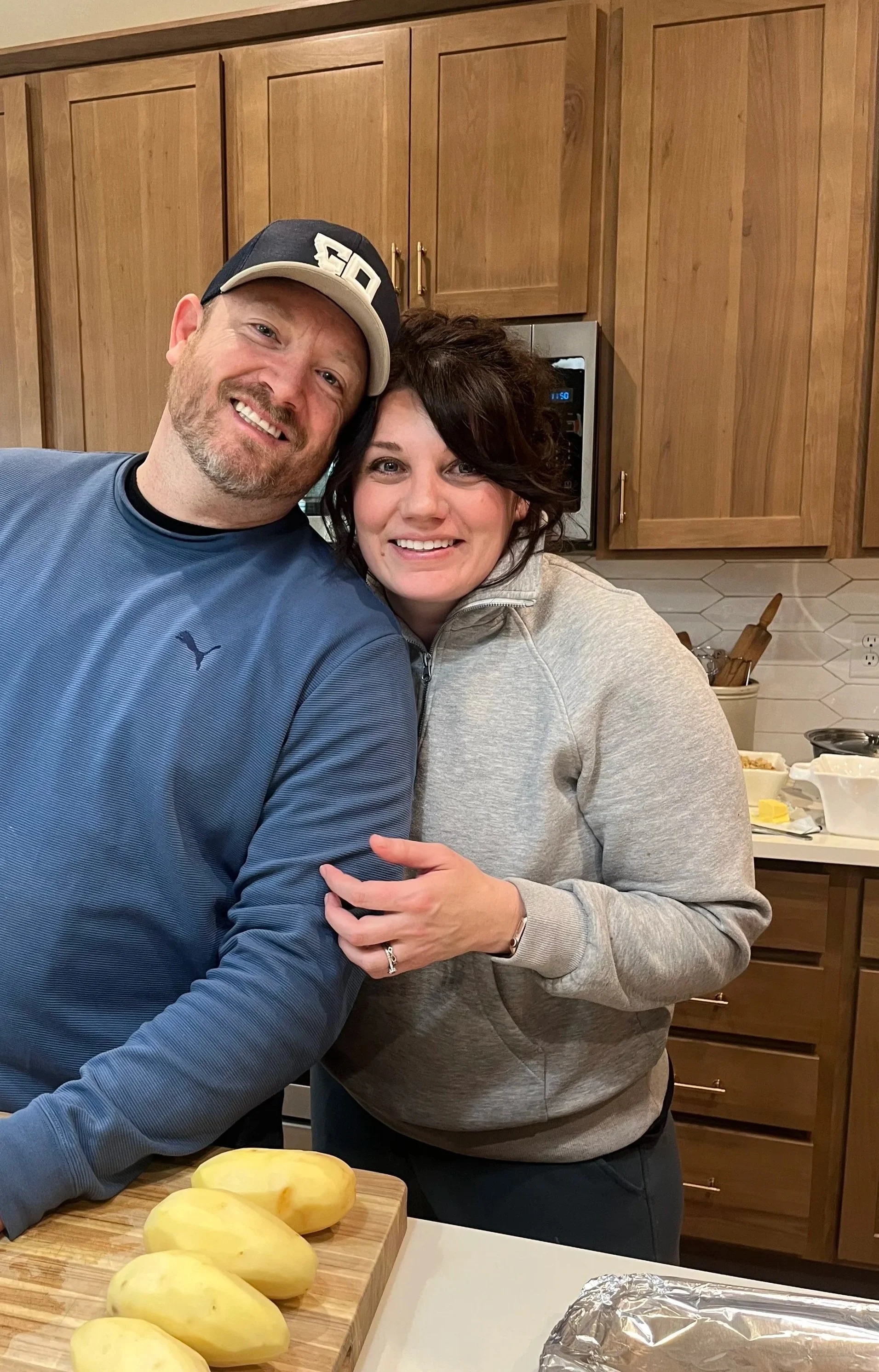 A smiling man and woman hugging in a kitchen, with peeled potatoes on a cutting board and foil on a baking tray in the foreground.