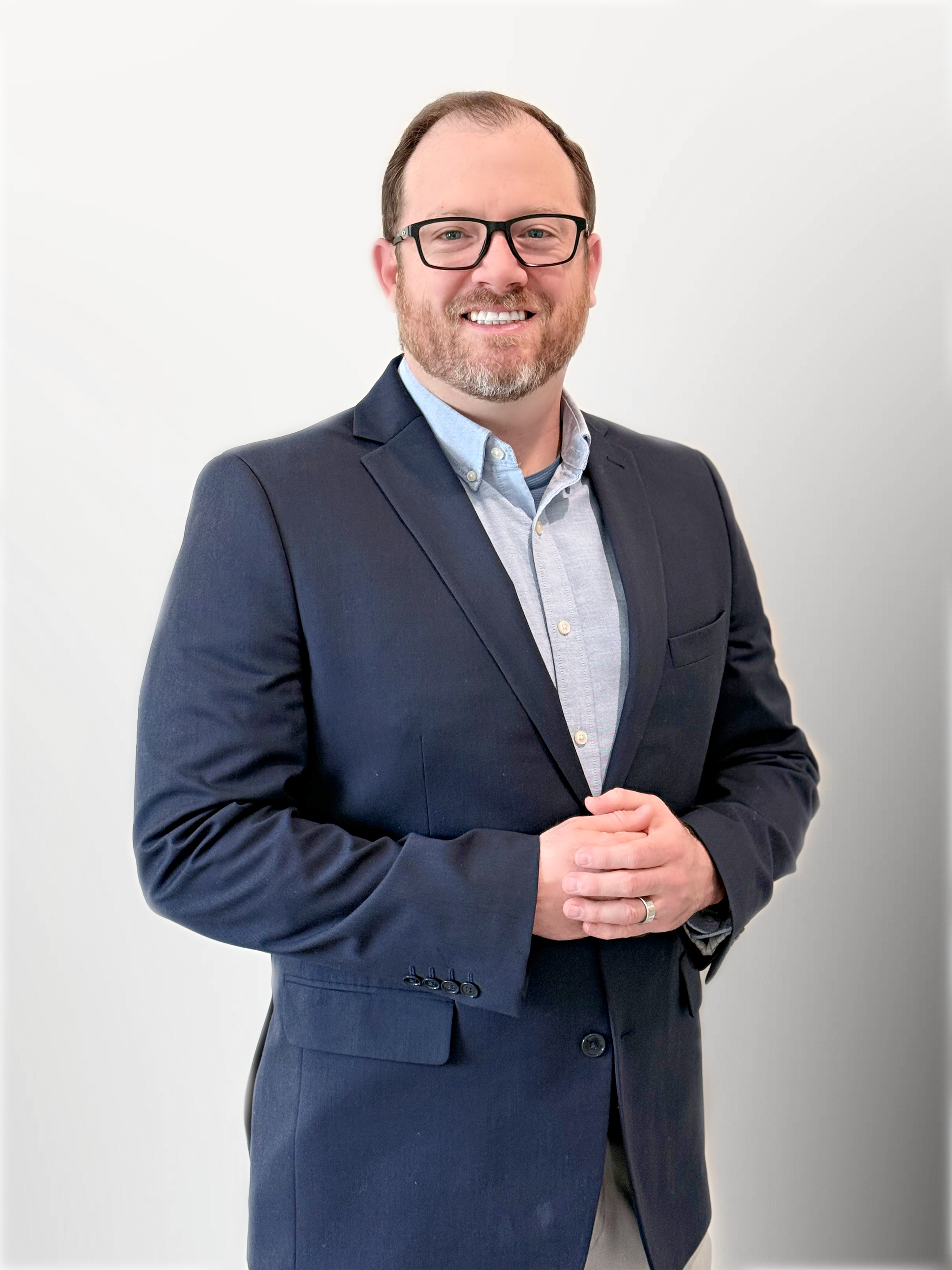 A man with glasses and a beard is smiling and dressed in a dark blue suit and light blue shirt, standing against a plain white background.