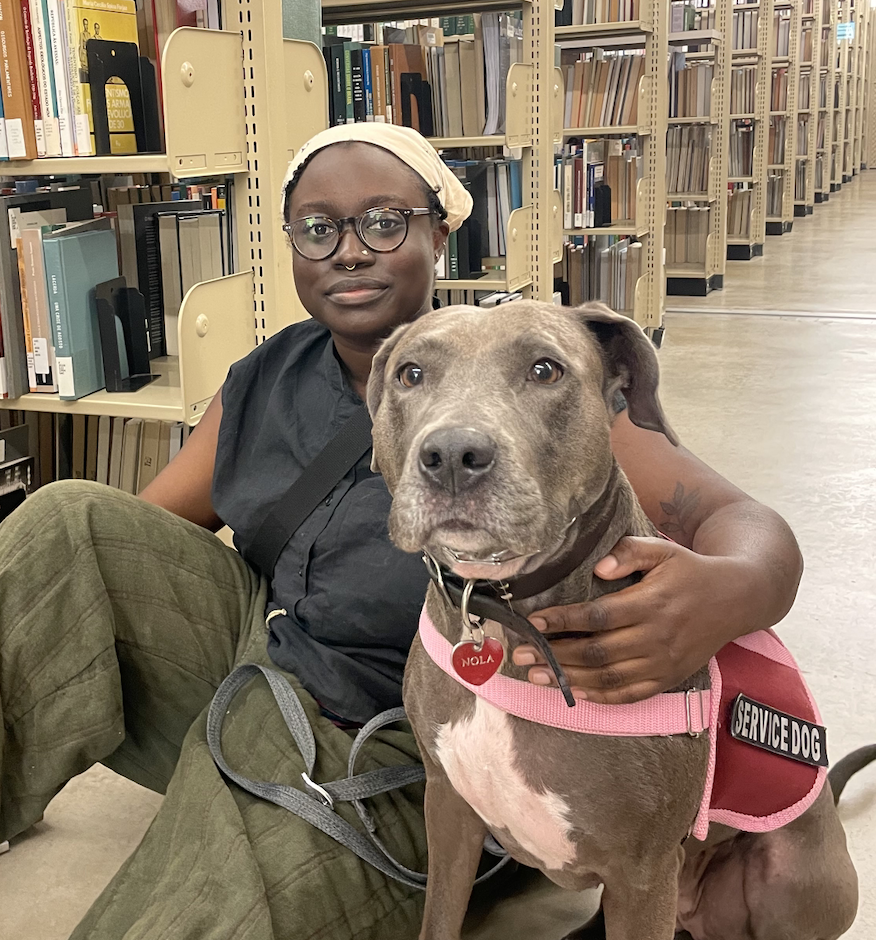 Image description: A photo of Suzanne and Nola sitting on the floor in the library. Suzanne wears a black shirt and green pants. Nola is a gray dog wearing a pink service dog vest. Suzanne has her arm around Nola. End description.