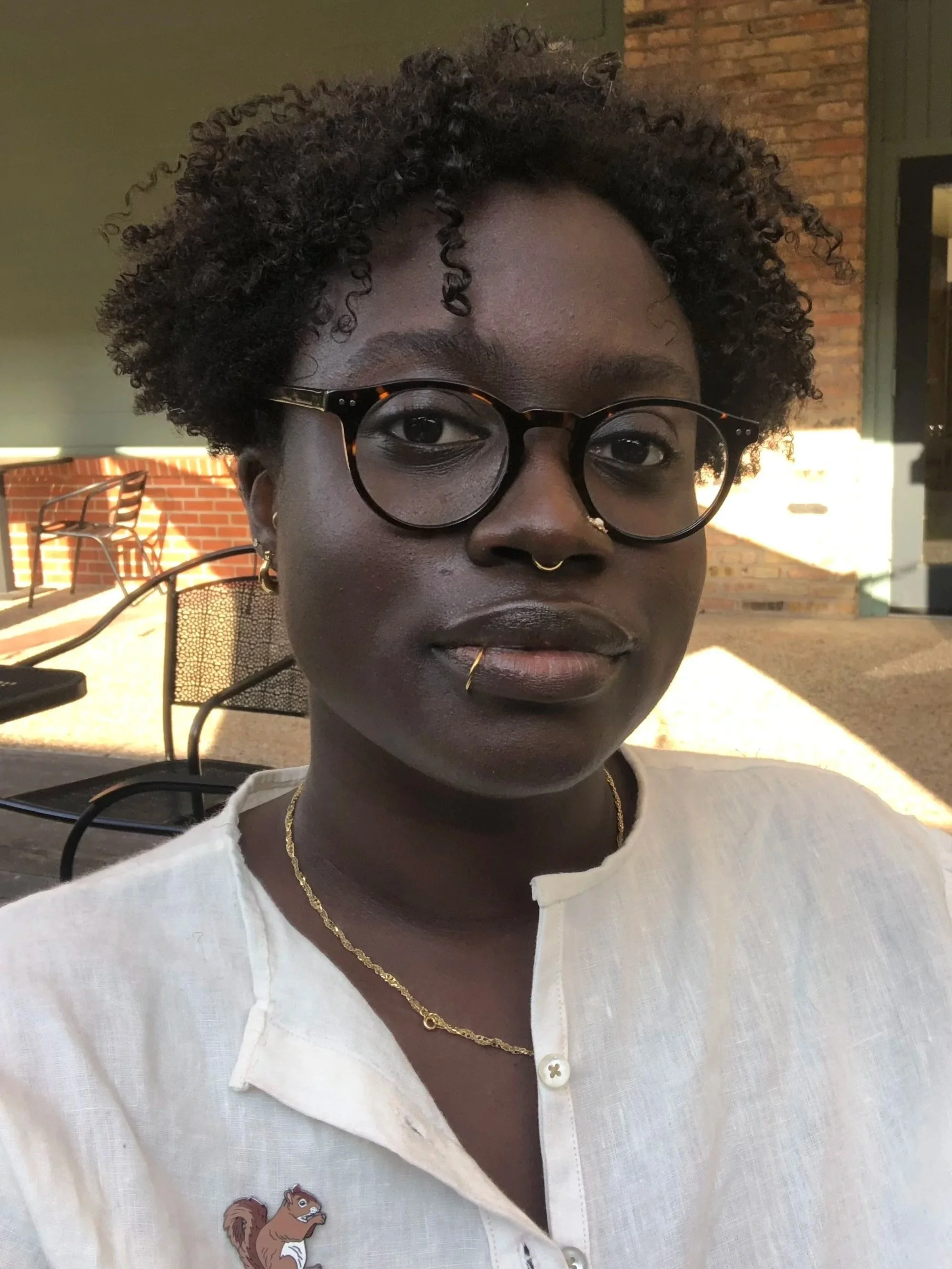 mage description: A photo of Suzanne sitting outside. Suzanne is Black and wears eyeglasses and gold jewelry, and is wearing white shirt with a squirrel pin. End description.
