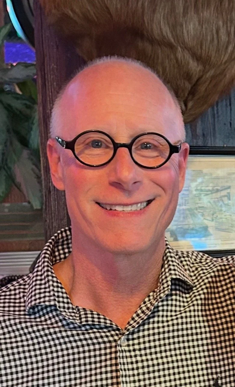 A smiling man with light skin, wearing round black glasses and a black-and-white checkered shirt, sitting indoors.