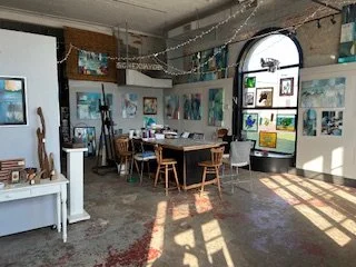 An art gallery with framed paintings and photographs on the walls, a table with chairs, and sunlight streaming through a large arched window.