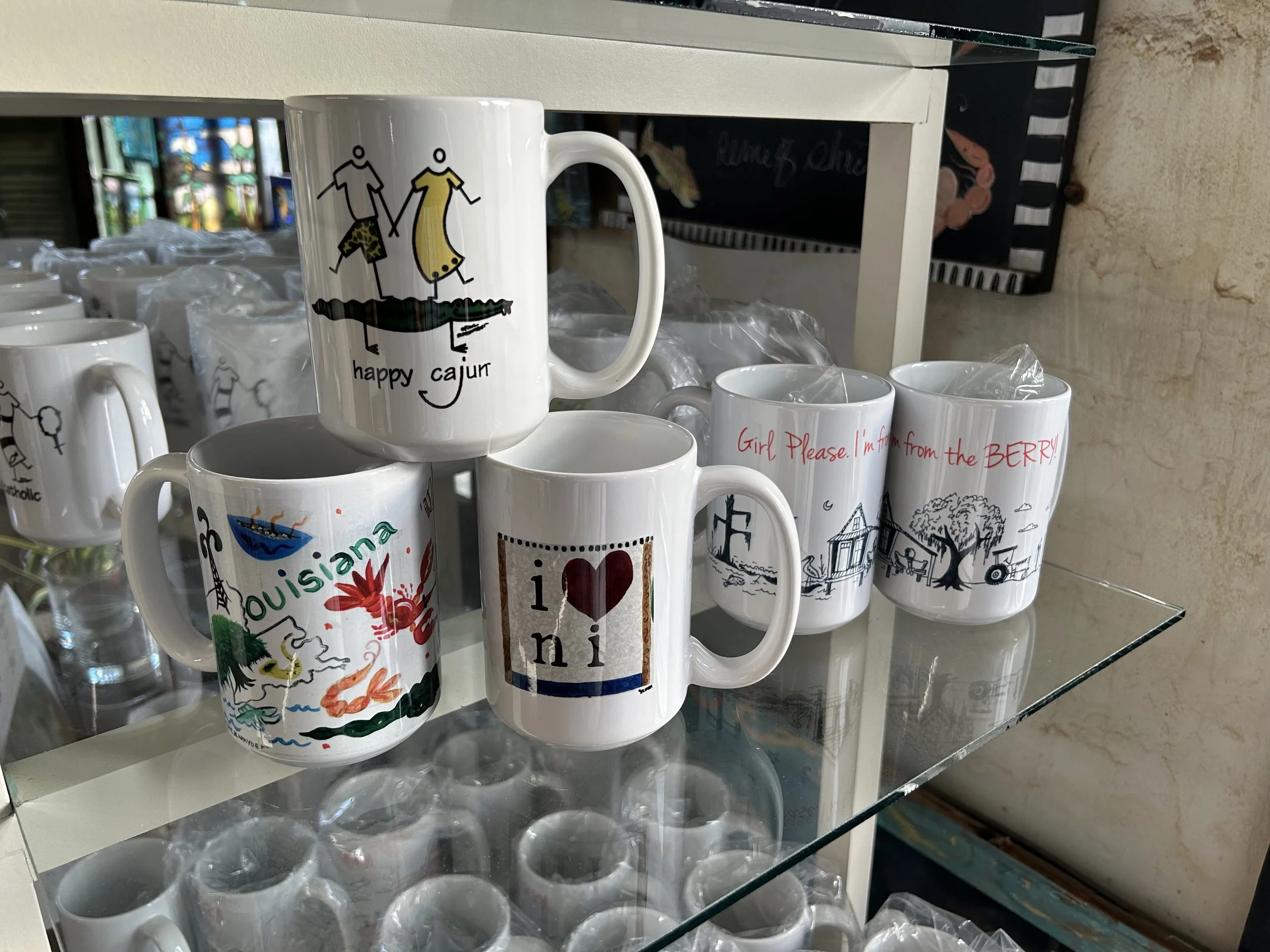 A display of white ceramic mugs with various colorful and black printed designs, focusing on a mug featuring a drawing of two stick-figure people holding hands, with the words "happy cajun" underneath, and others with different graphics and text, arranged on glass shelves.