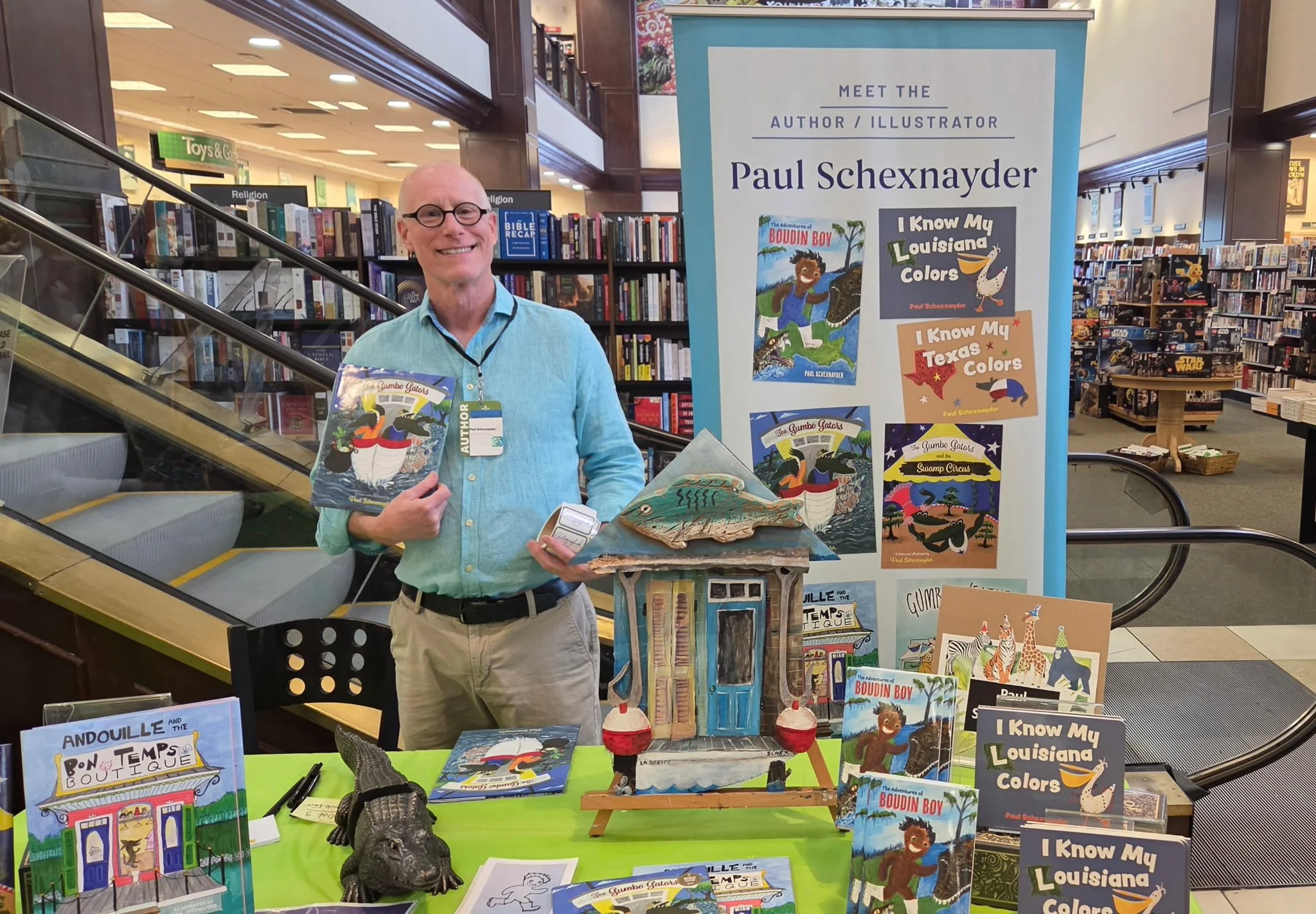 A man with glasses and a light blue shirt standing at a book signing booth in a bookstore, holding a children's book titled 'The Adventures of Boudin Boy.' The booth displays various colorful children's books by author Paul Schexnayder, including titles like 'I Know My Louisiana Colors' and 'I Know My Texas Colors.' Behind him are bookshelves filled with books and a large vertical sign introducing him as the author and illustrator.
