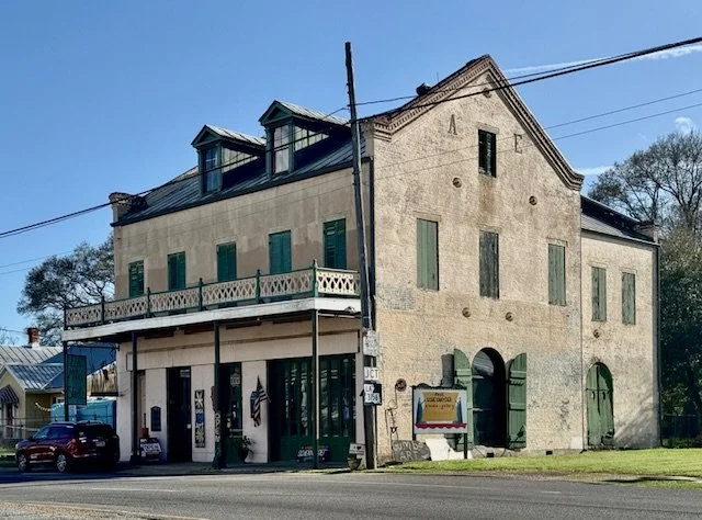 An old, multi-story brick building with boarded-up windows, green shutters, and a corner entrance, situated along a street with parked cars and a utility pole in front.