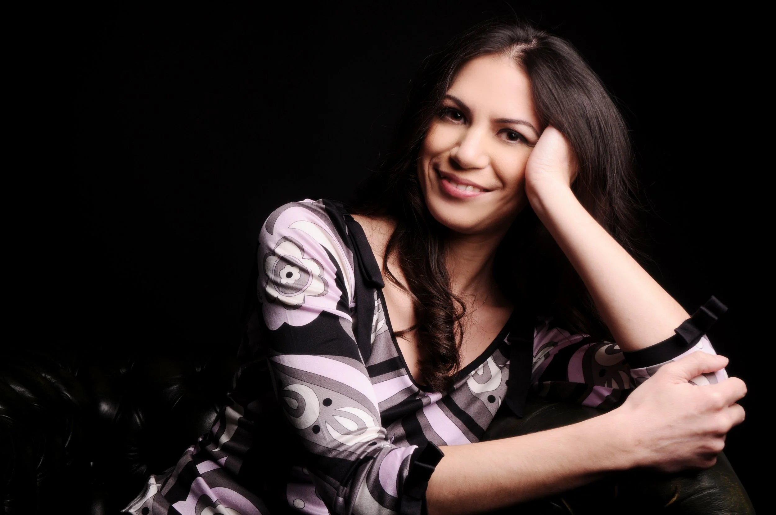 A woman with long dark hair smiling and resting her head on her hand, wearing a black and white patterned top, sitting on a black leather couch against a black background.