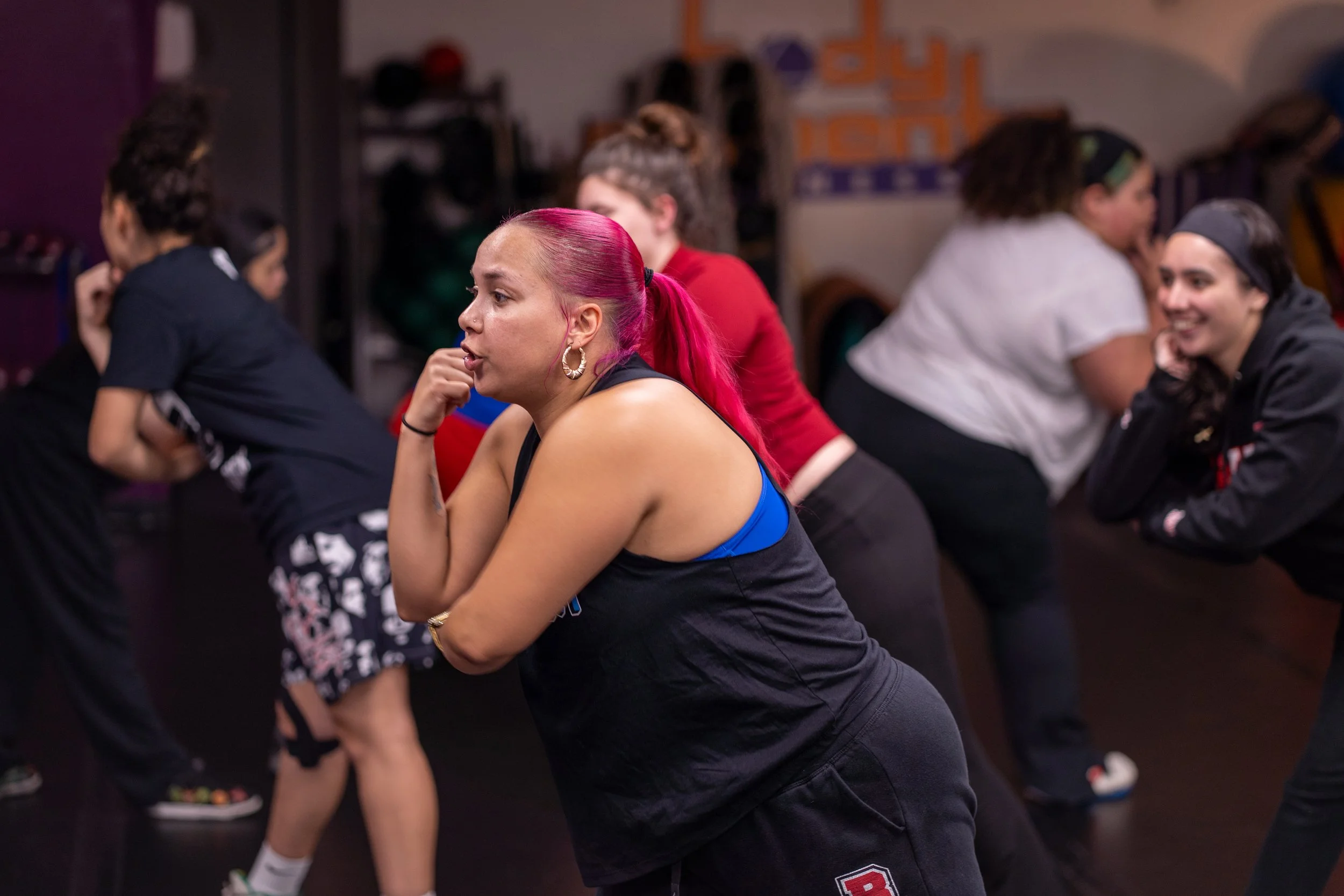 A woman with bright pink hair tied in a ponytail participates in a fitness class, surrounded by other women in workout attire in a gym or exercise studio.