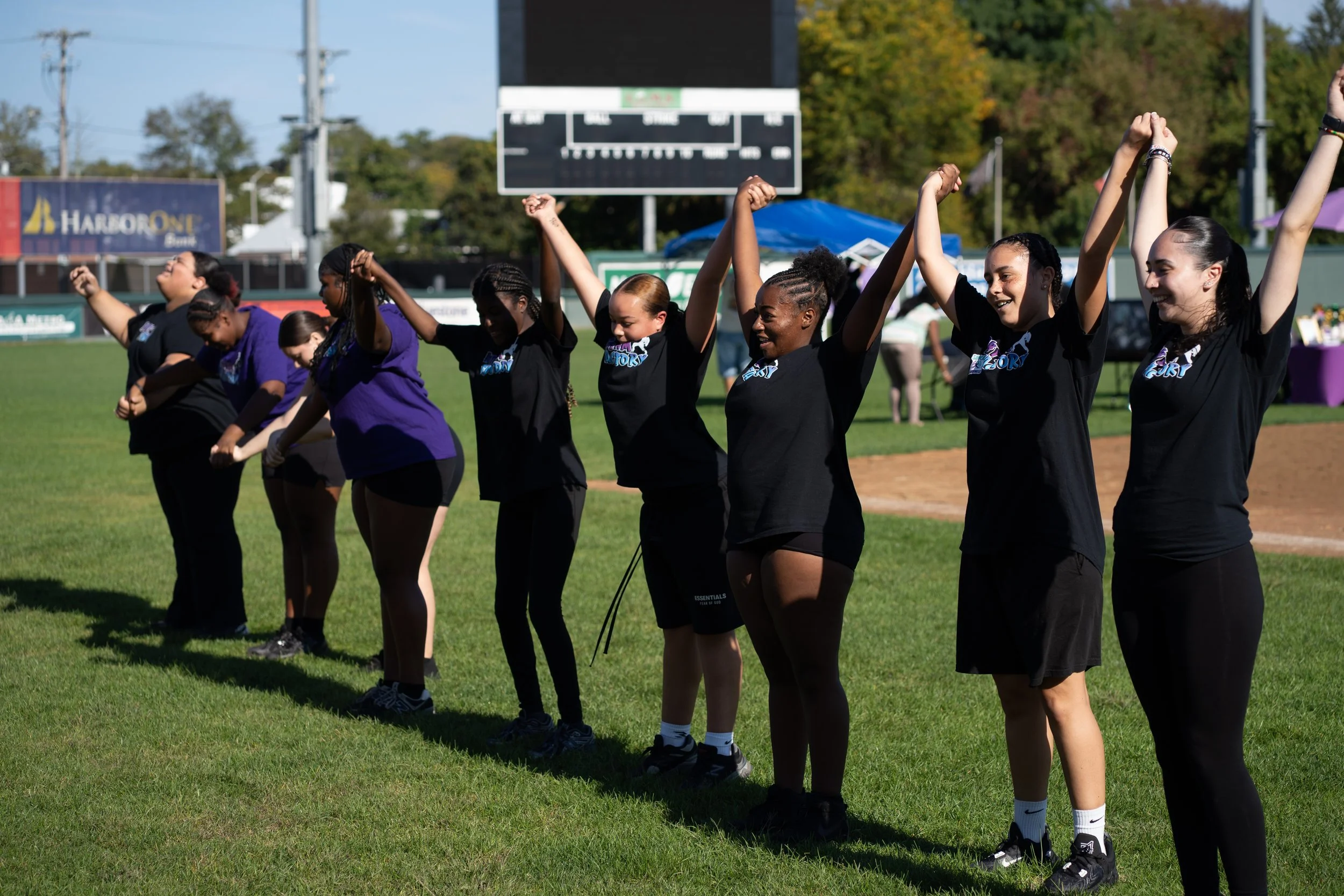 A group of diverse women standing in a line on a grassy field, holding hands and raising their arms in celebration or solidarity at a sports event, with trees, tents, and a scoreboard in the background.