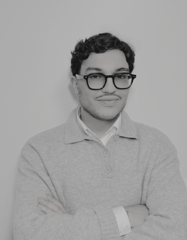 Jeune homme portant des lunettes épaisses, souriant légèrement, en chemise blanche et pull gris, avec les bras croisés.