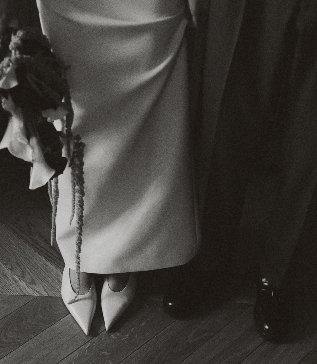 Chaussures à talons pointus en cuir clair, une personne portant une longue jupe ou robe sombre, un bouquet de fleurs, et un sol en parquet.