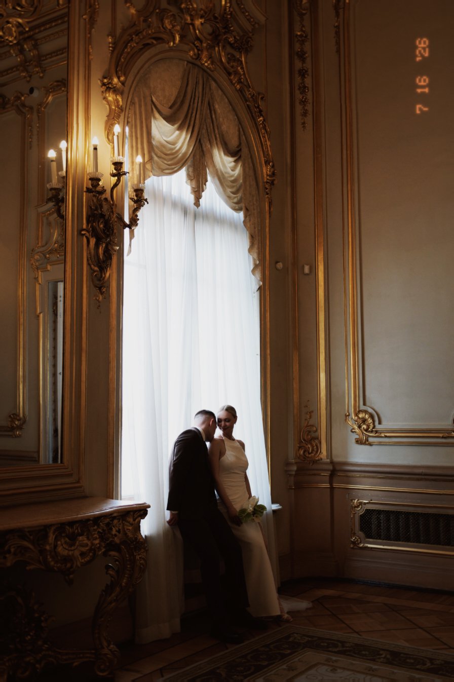 Un couple en costume de mariage dans une pièce ornée de dorures, de grands rideaux blancs et de murs décorés, dans une ambiance élégante.