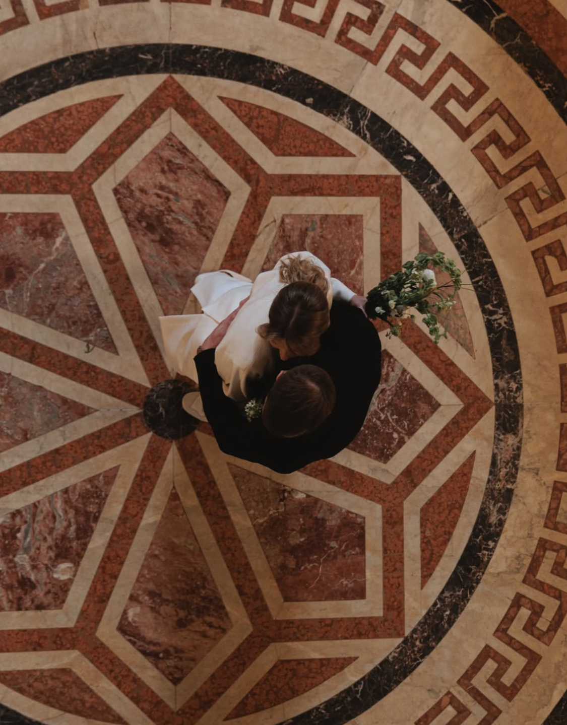 Un couple dans une robe blanche de mariage et un costume noir qui dansent sur un sol en marbre décoré, vu du dessus, au centre d'un motif géométrique.