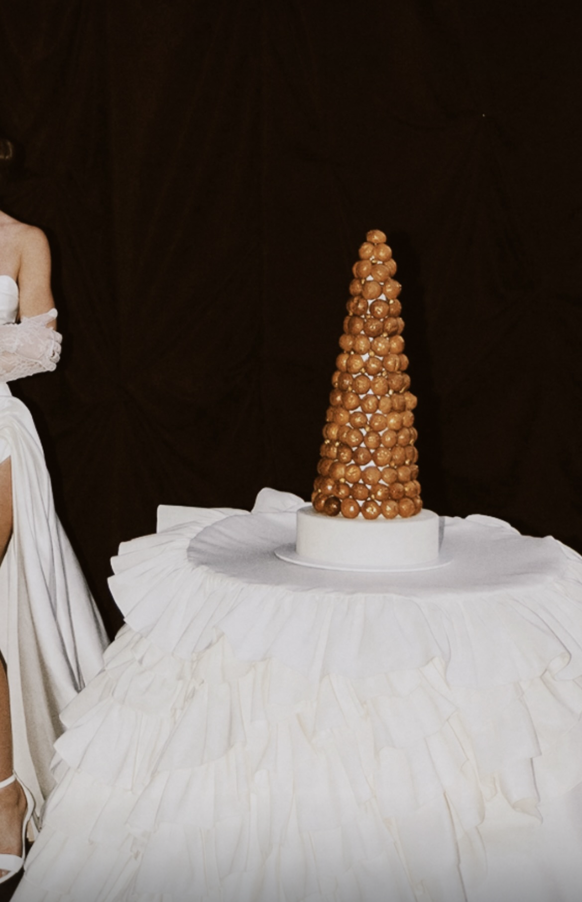 Un gâteau en forme de tour de macarons marrons, placé sur une table avec une nappe blanche. À gauche, partie d'une personne en robe blanche.