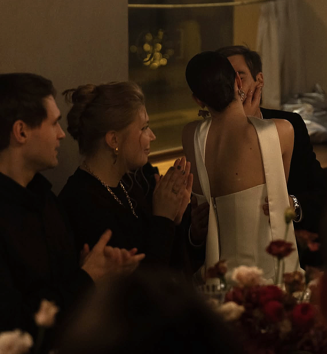 Une scène de mariage où une femme en robe blanche embrasse un homme en costume noir, entourée de personnes qui applaudissent et regardent l'événement.