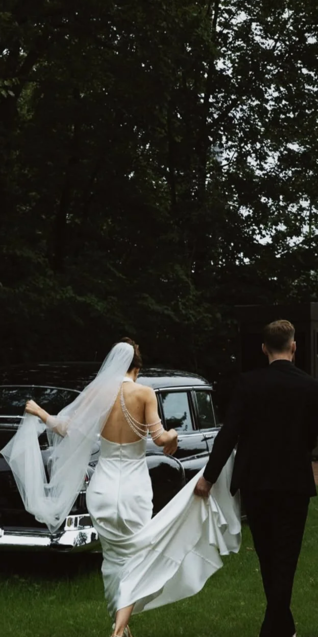 Un couple en tenue de mariage : la mariée en robe blanche et un voile, le marié en costume noir, près d'une voiture noire dans un environnement naturel.