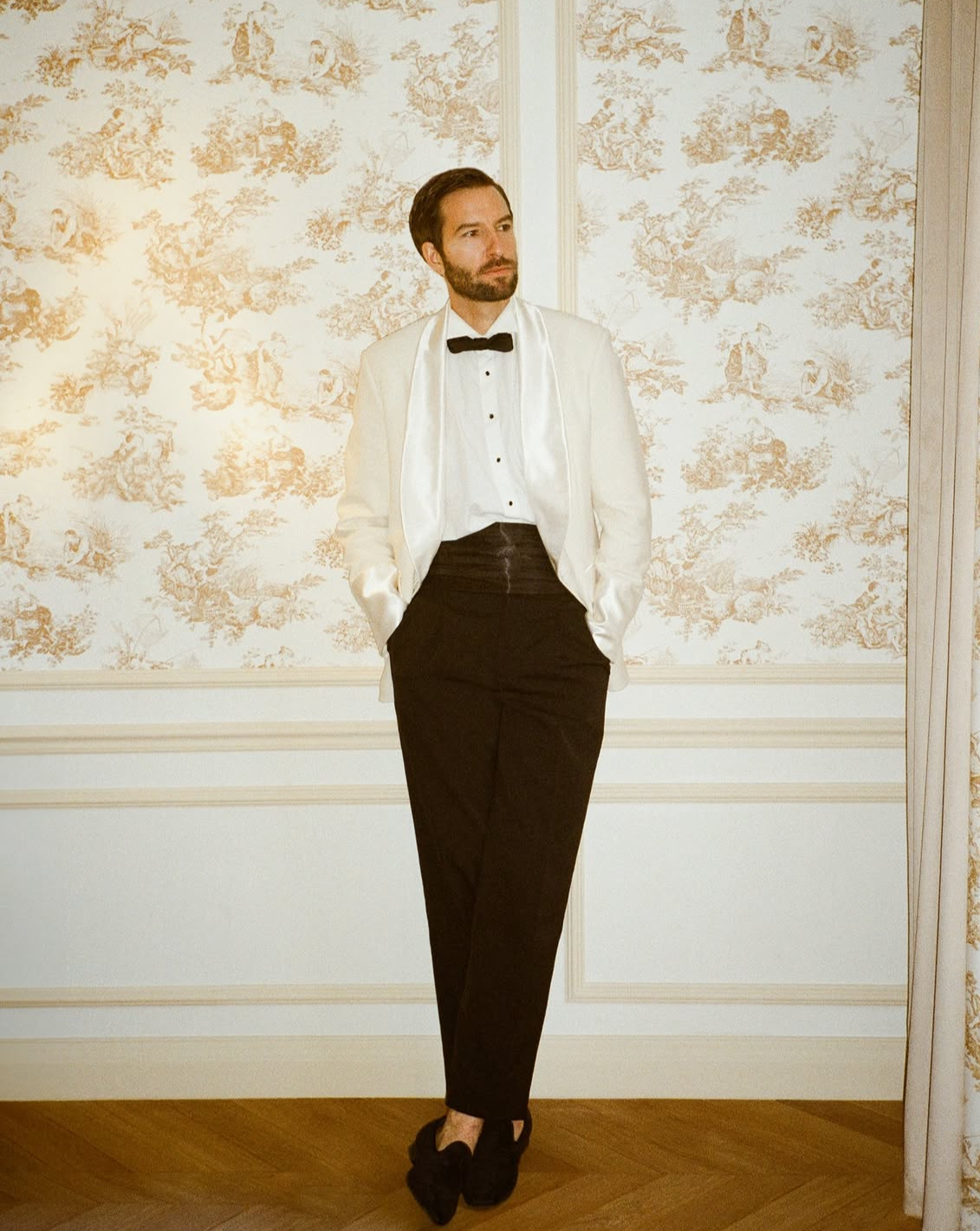 Un homme habillé en smoking noir avec une veste blanche, une chemise blanche, une cravate noire, et des chaussures noires, posant dans une pièce ornée de papier peint avec un motif floral.