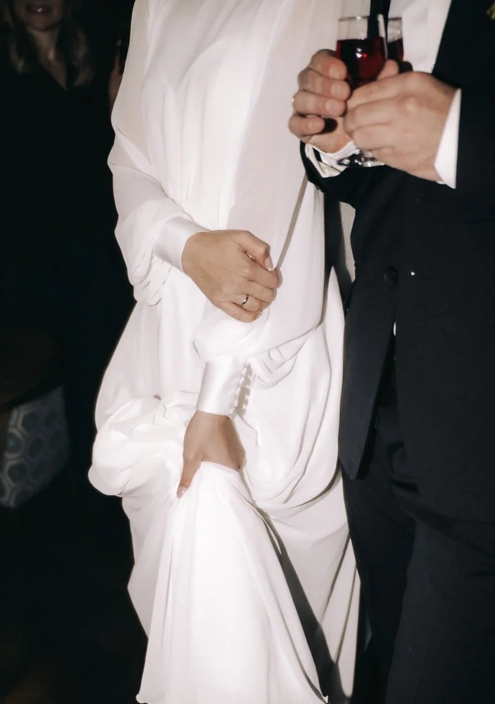 Une personne en robe blanche tient la main d'une autre personne en costume noir qui tient un verre de vin rouge.