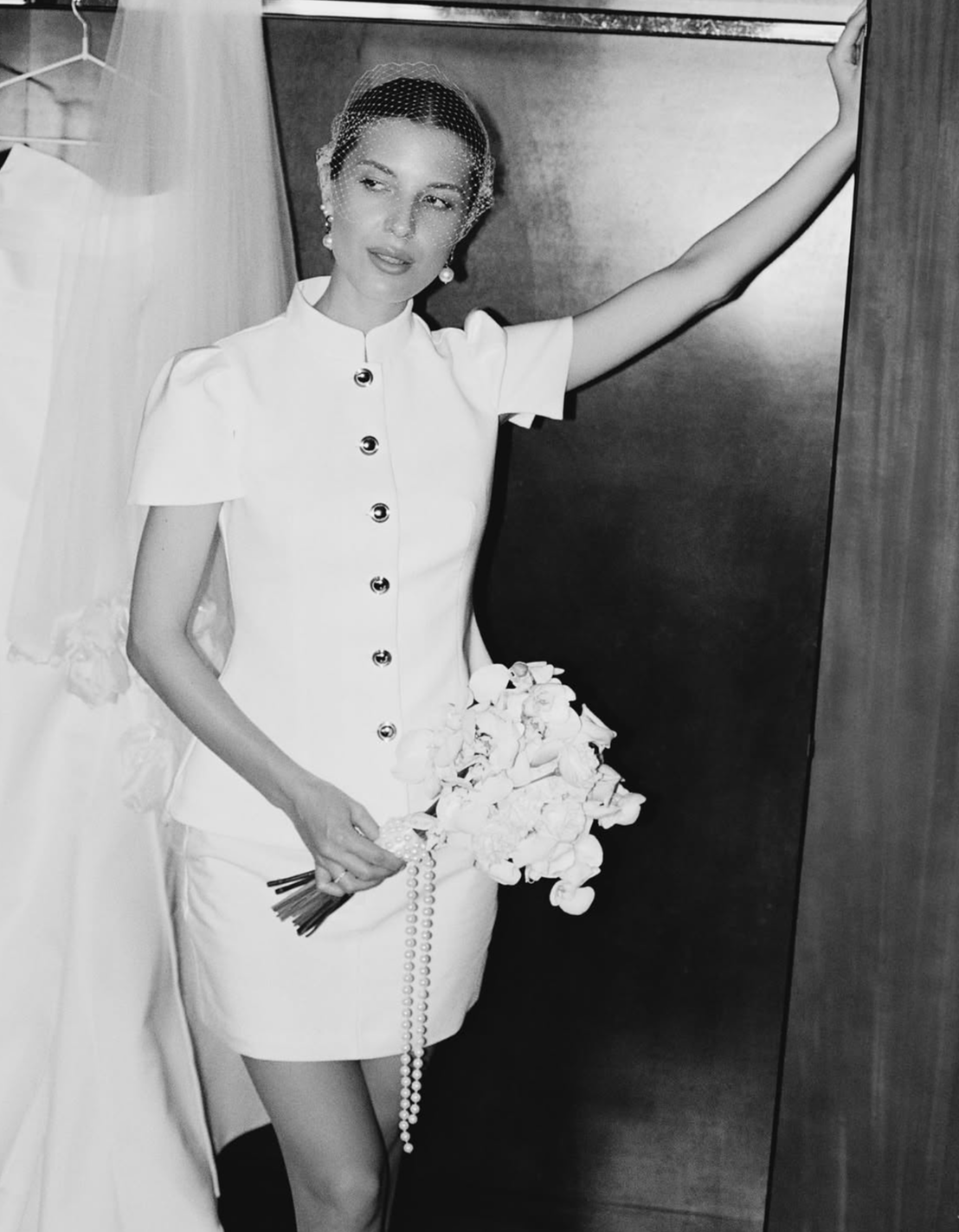 Une femme élégante portant une robe blanche, tenant un bouquet de fleurs, se tient près d'une porte dans un décor intérieur.