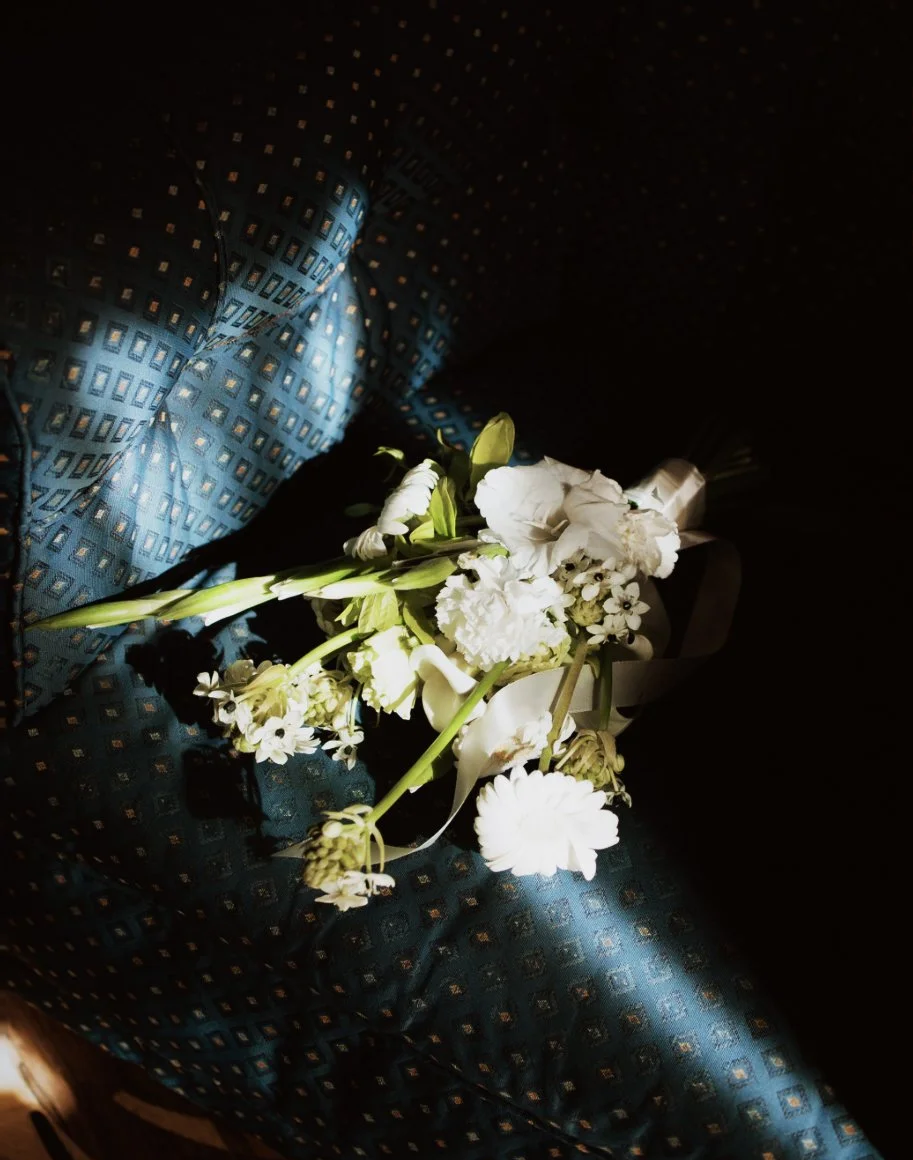 Un bouquet de fleurs blanches sur un lit de tissu bleu avec un motif géométrique, éclairé par une lumière directe, dans un environnement sombre.