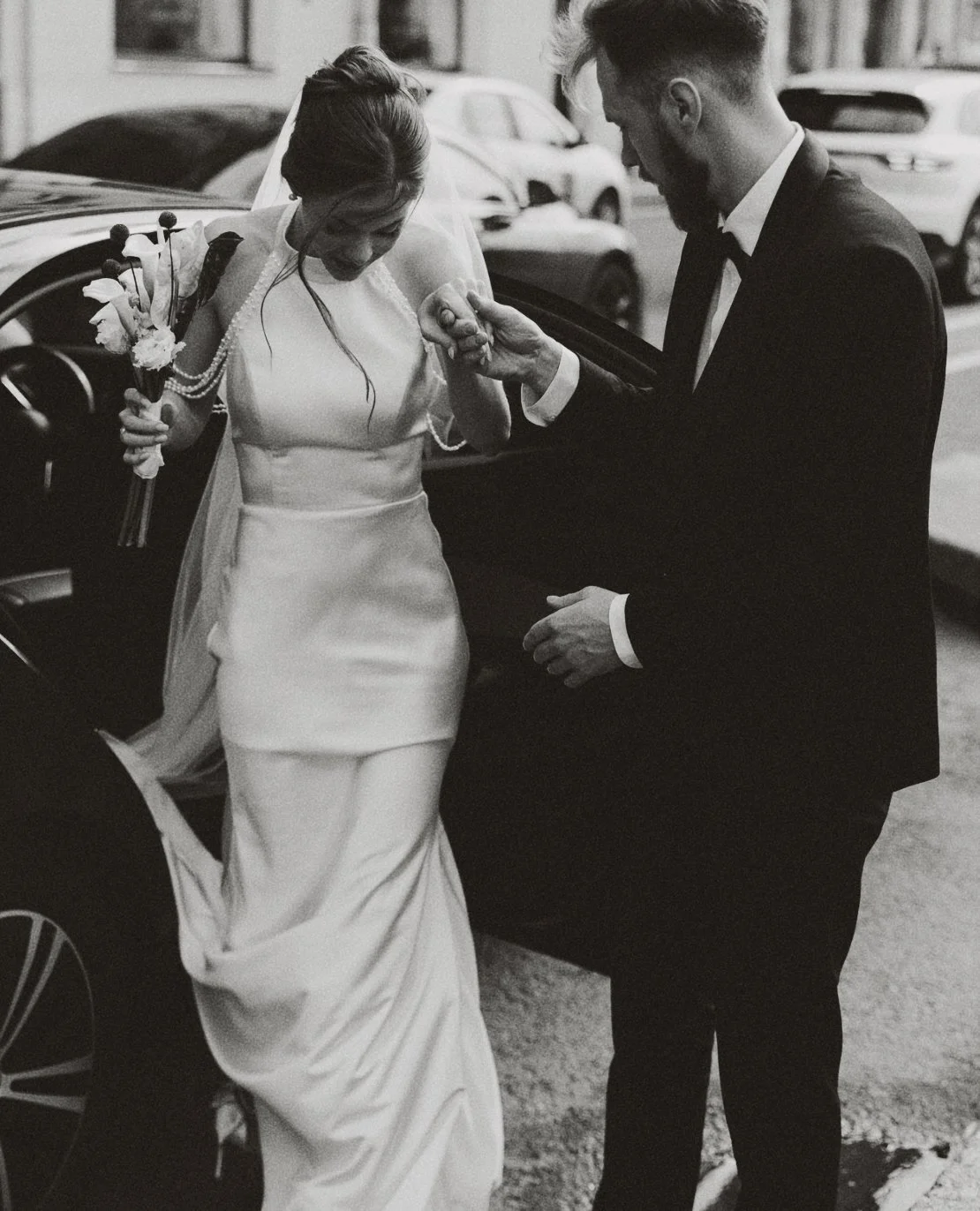 Un couple en vêtements de mariage, la femme portant une robe de mariage et tenant un bouquet, un homme en costume noir, interagissent devant une voiture.