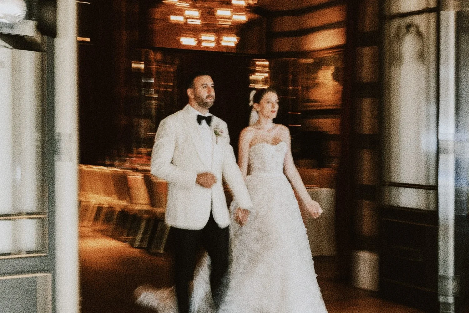 Un couple en habits de mariage, un homme en smoking blanc avec nœud papillon et une femme en robe de mariée blanche, marchant dans une salle élégante avec des lumières tamisées.