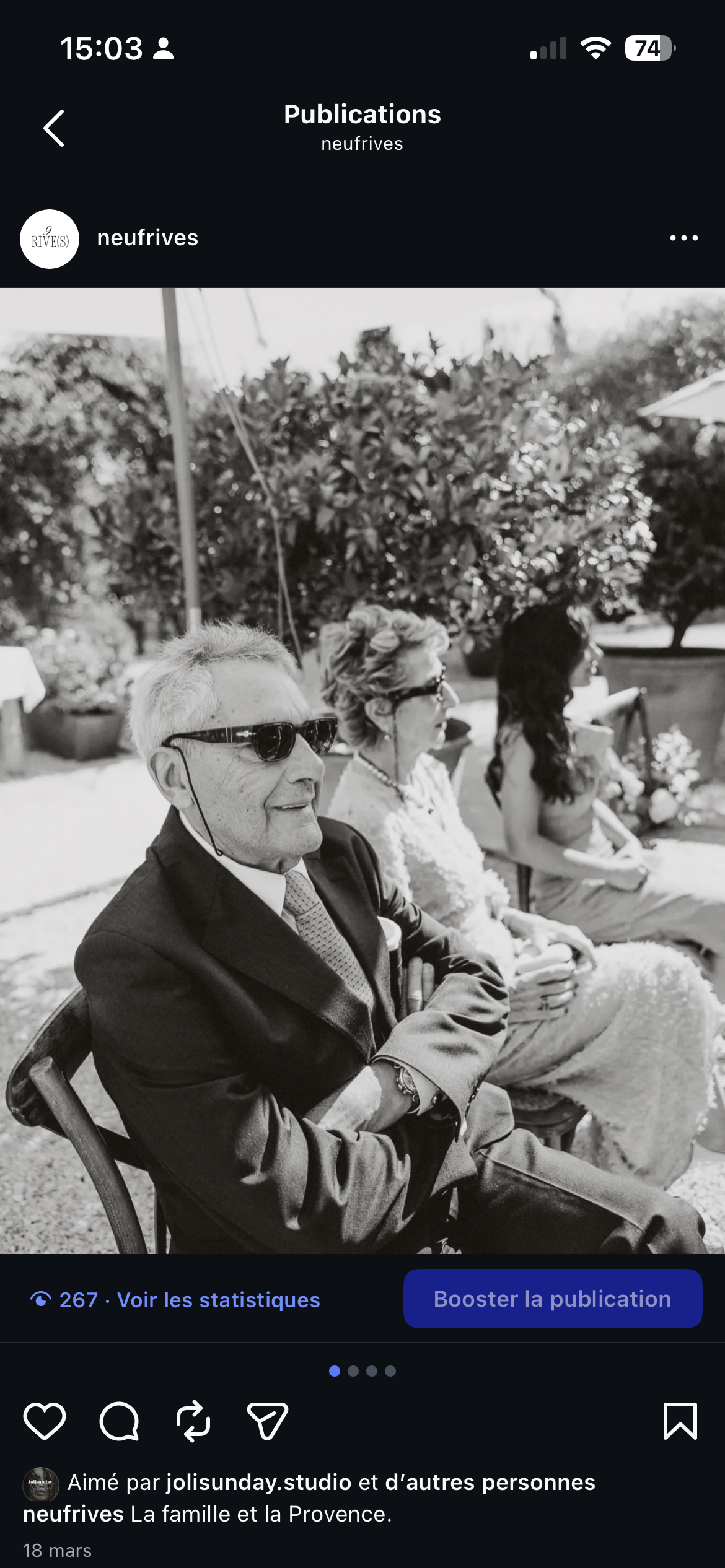 Photo en noir et blanc d'une famille assise en extérieur, probablement lors d'un événement, avec un homme en costume portant des lunettes de soleil, une femme en robe élégante et une autre femme. Arrière-plan avec des arbres et des plantes.